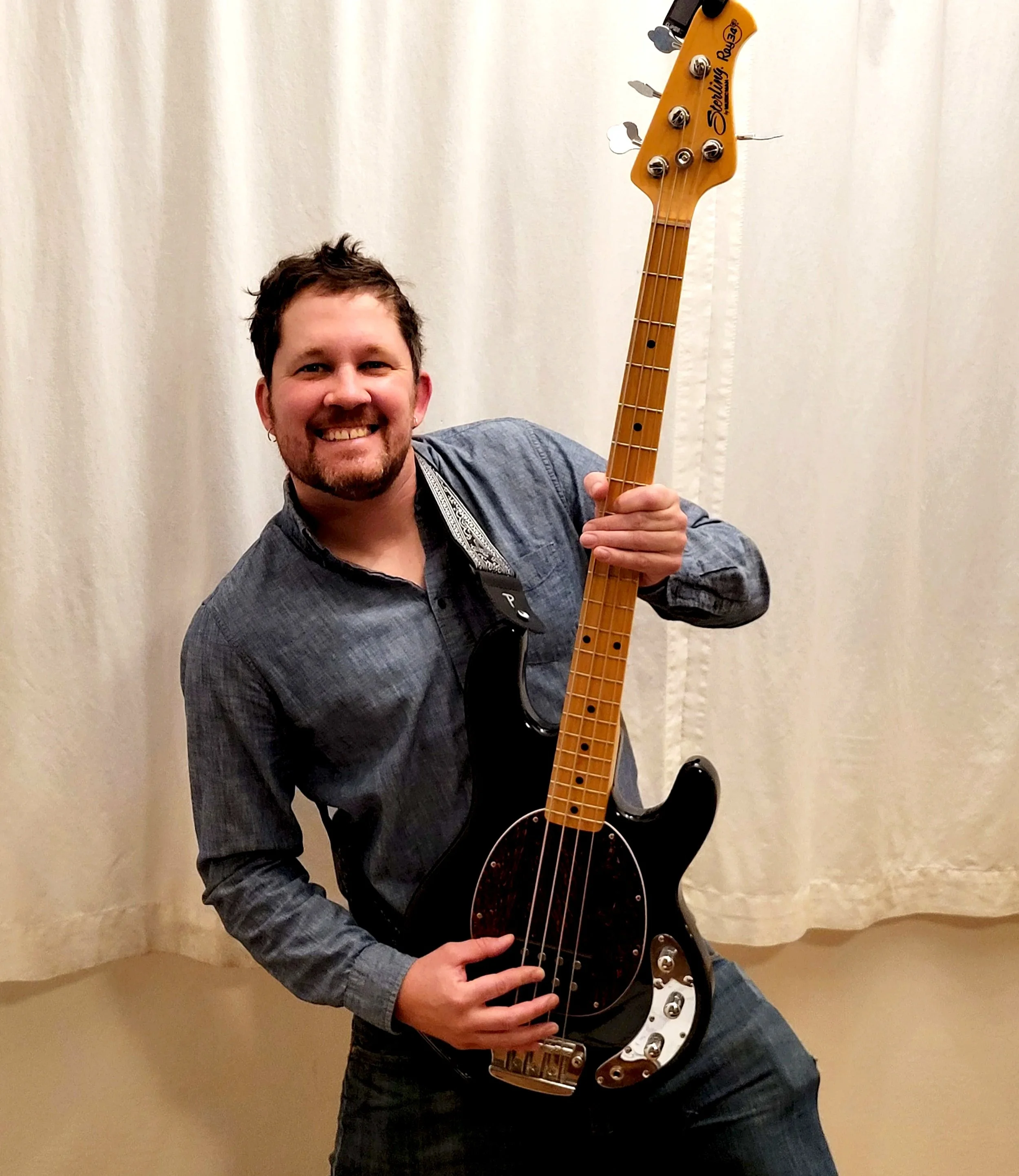 A man holding a black and brown electric bass guitar, smiling, in front of a beige curtain.