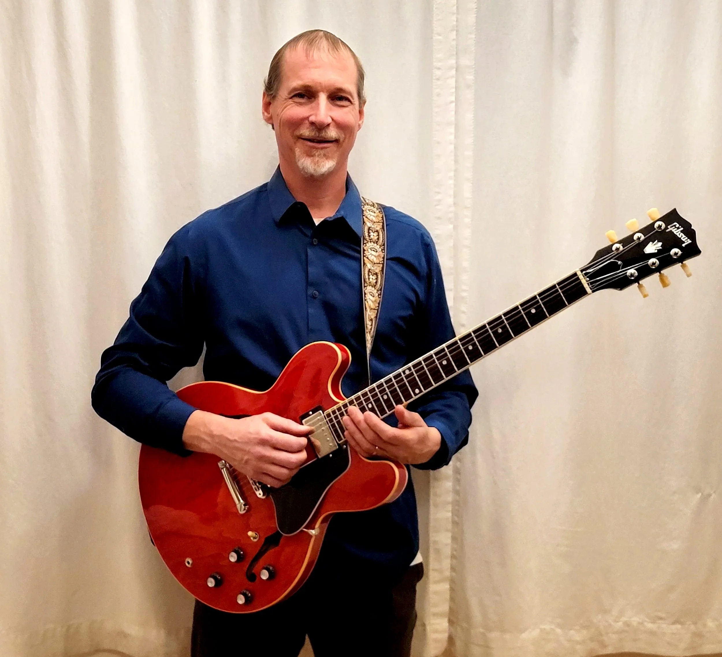 A man in a blue shirt holding a red Gibson electric guitar, standing in front of a cream-colored curtain.