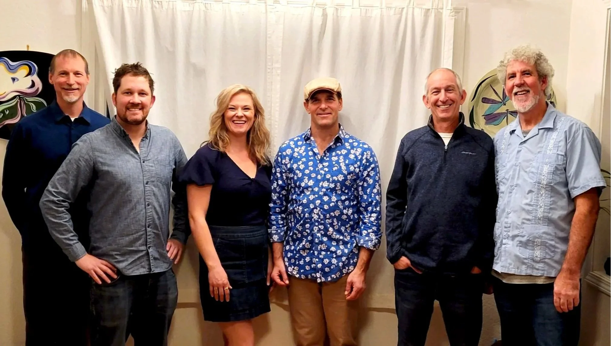 A group of six people standing indoors, smiling at the camera, with a white curtain behind them and colorful artwork on the walls.