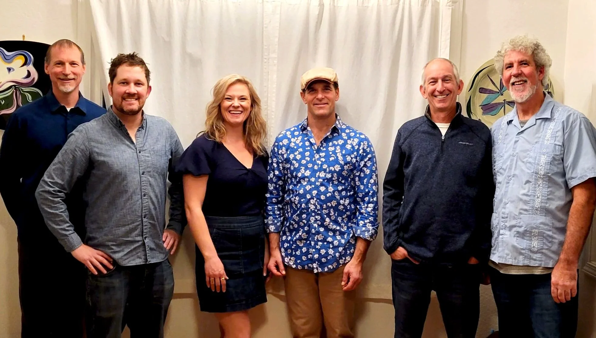 Group of seven people standing indoors against a white curtain, smiling at the camera.