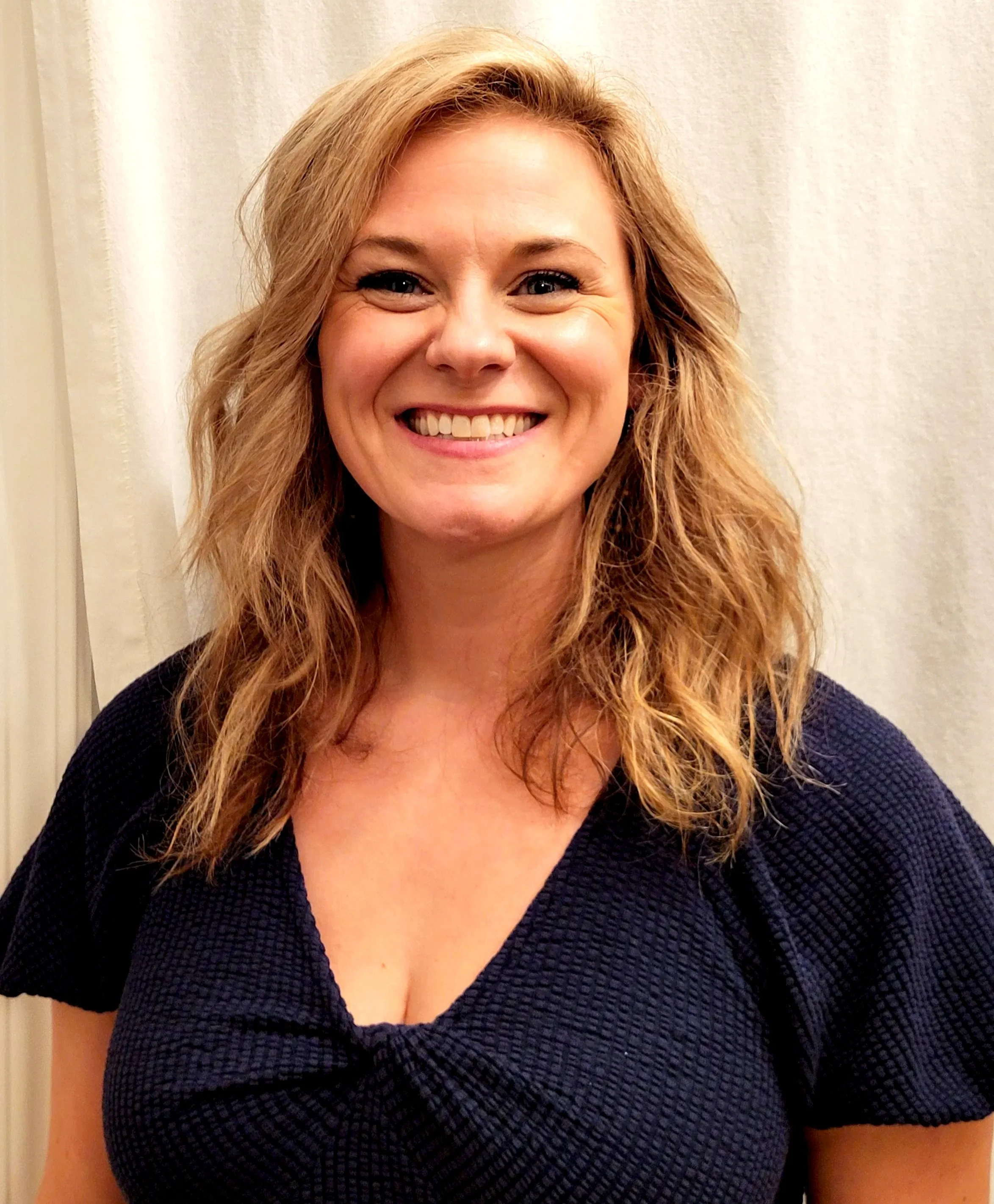 A woman with shoulder-length wavy blonde hair, smiling, wearing a black textured top, standing in front of a light-colored curtain.