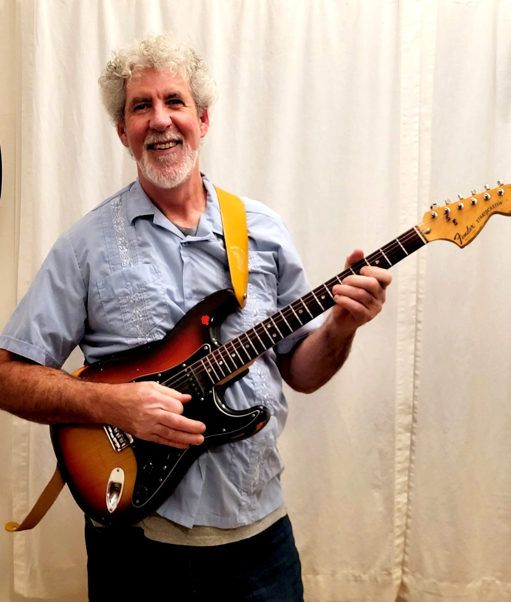 A smiling man with gray curly hair and beard playing an electric guitar.