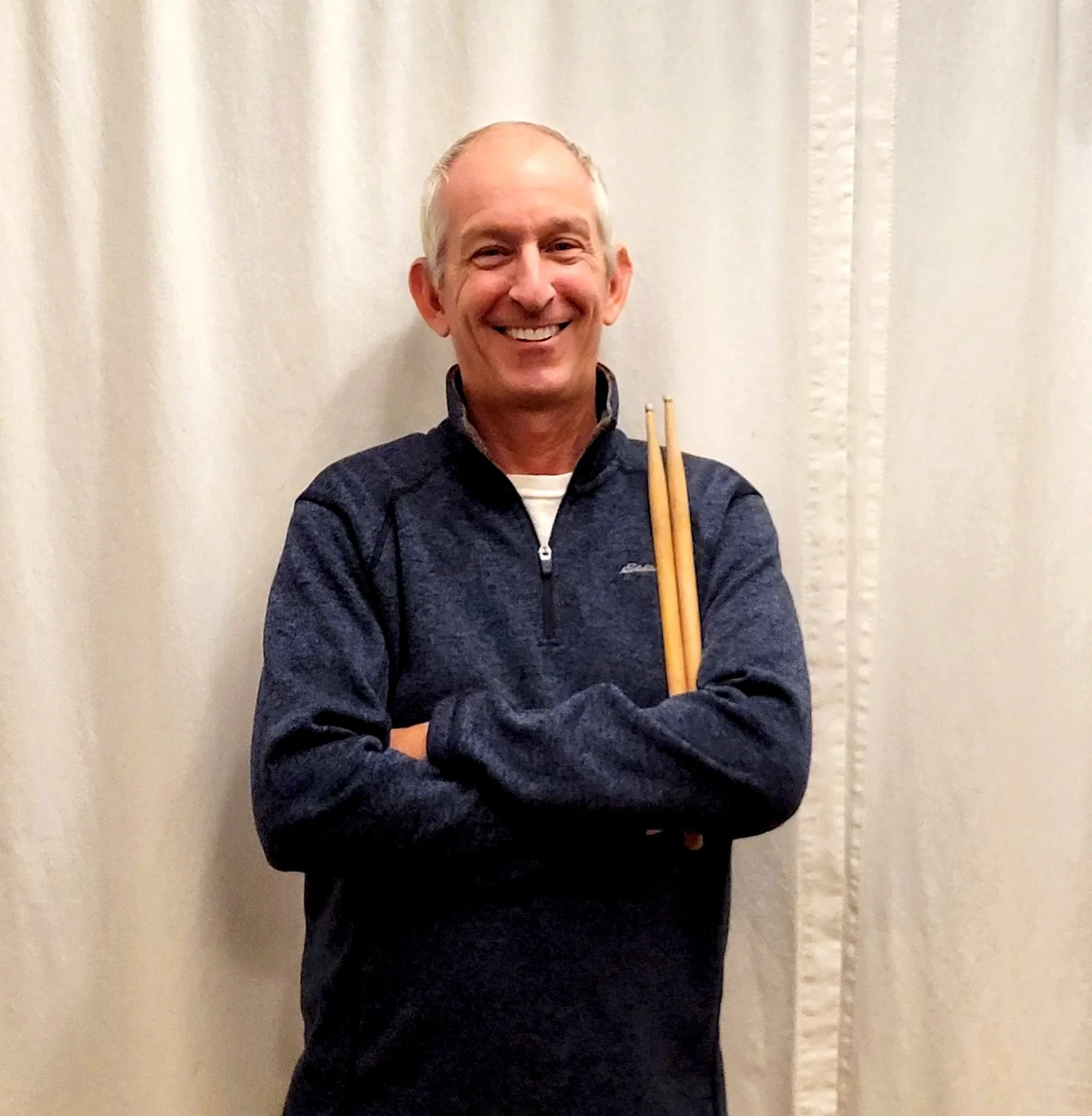 A smiling man with short, gray hair standing in front of a beige curtain, holding a pair of drumsticks across his chest.
