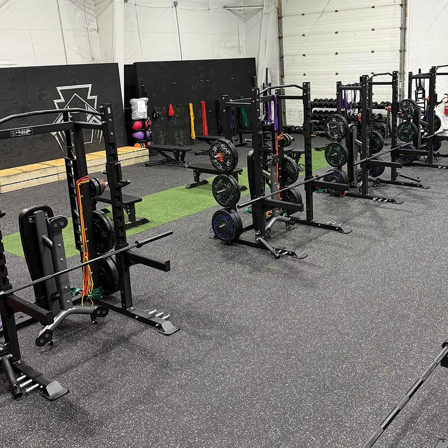 Empty gym with weightlifting equipment including barbells, plates, and resistance bands, on a speckled rubber floor with a green artificial turf strip.