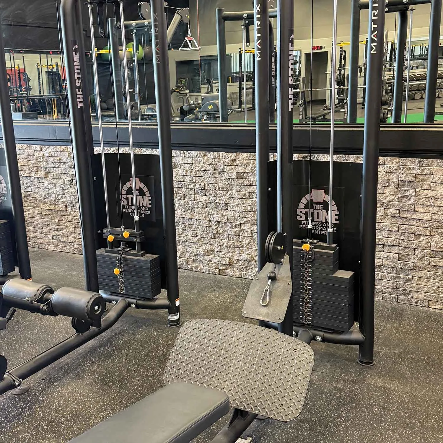 Inside a gym, a cable machine with weight stacks and a piece of workout equipment with a textured metal seat and padded bench, windows reflecting gym equipment.