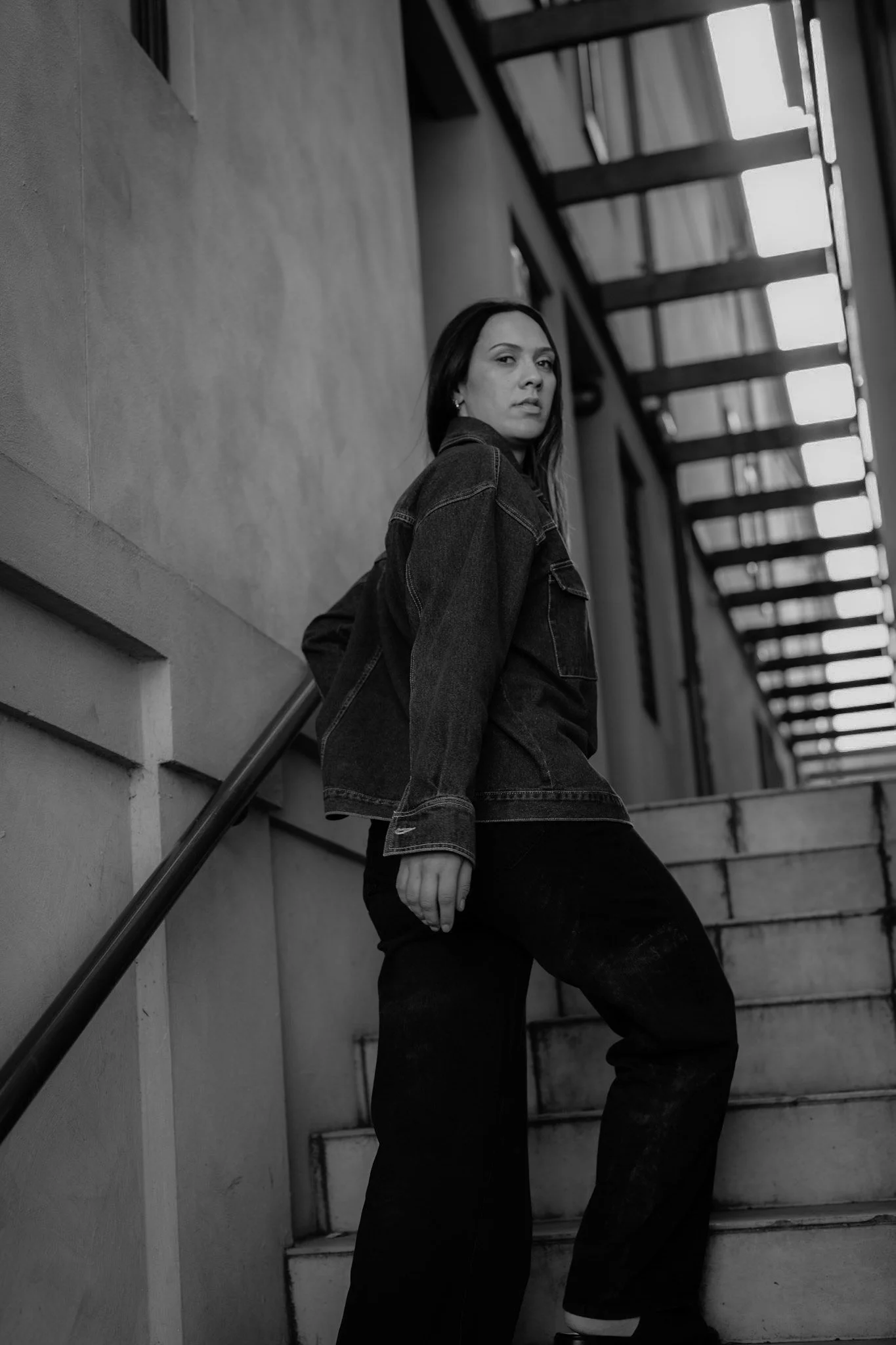 A woman in dark jeans and a denim jacket leaning against a staircase railing in an urban setting, looking at the camera with a serious expression.