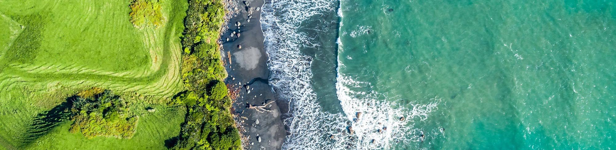 Aerial view of a coastline showing green grassy area on the left and ocean waves crashing onto a rocky beach on the right.