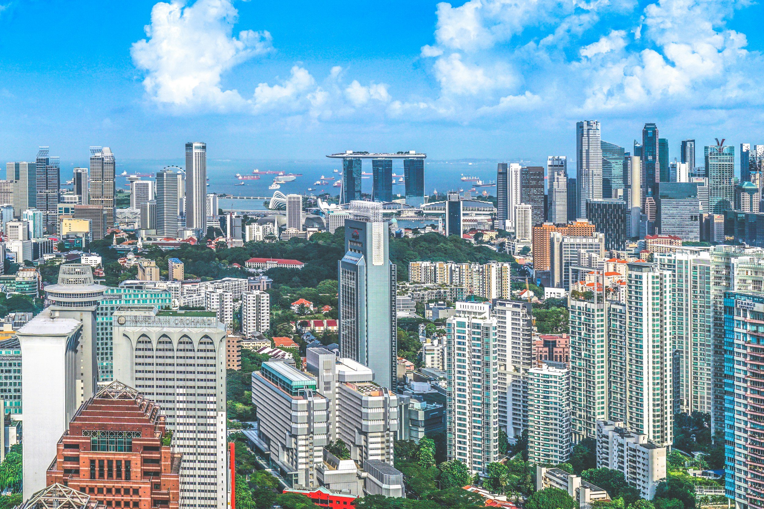 Aerial view of Singapore skyline with numerous skyscrapers, Marina Bay Sands hotel, and ships in the harbor under partly cloudy sky.