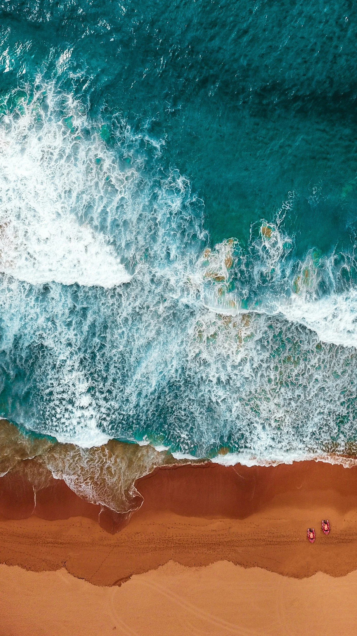 Aerial view of a beach with turquoise ocean waves crashing onto a sandy shore where two pink paddle boats are positioned.