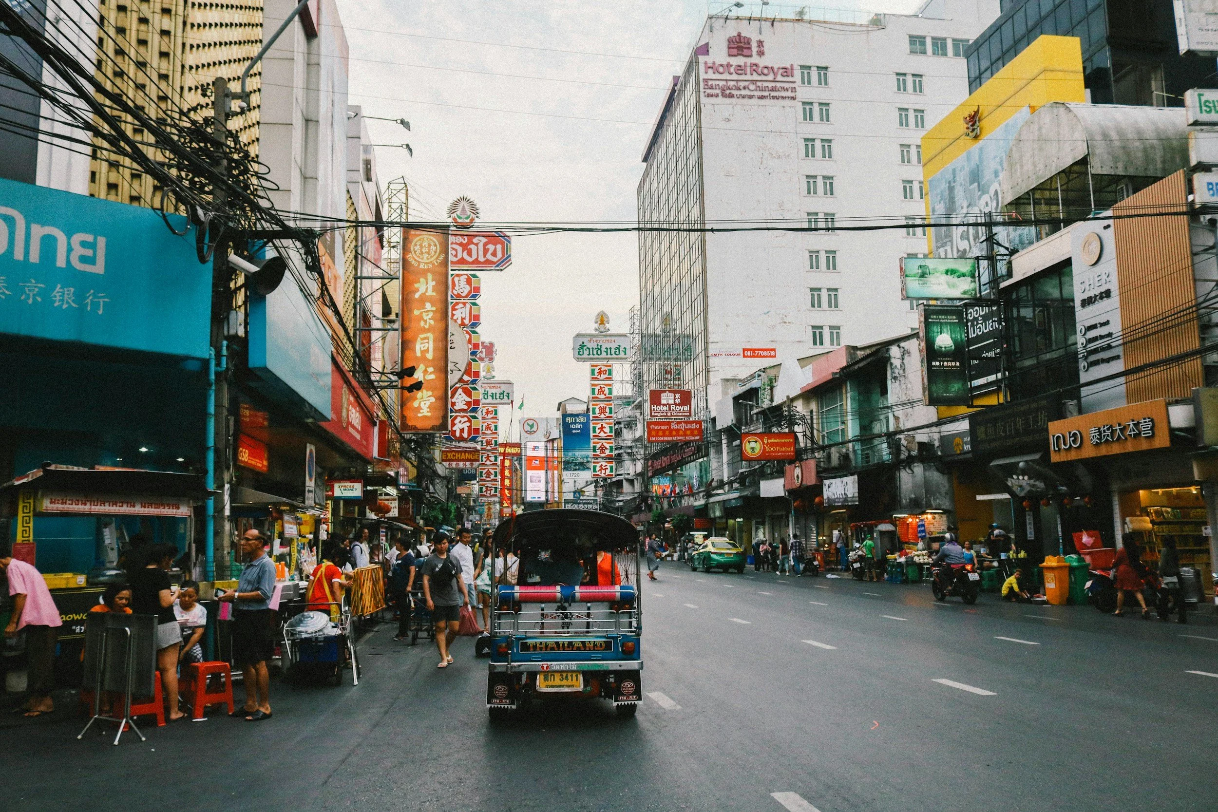 A busy city street in an Asian country, with various signs and advertisements in multiple languages, including Thai and Chinese. The street is filled with pedestrians, small shops, street vendors, and a tuk-tuk in the center. Tall buildings and colorful signage line both sides of the street.