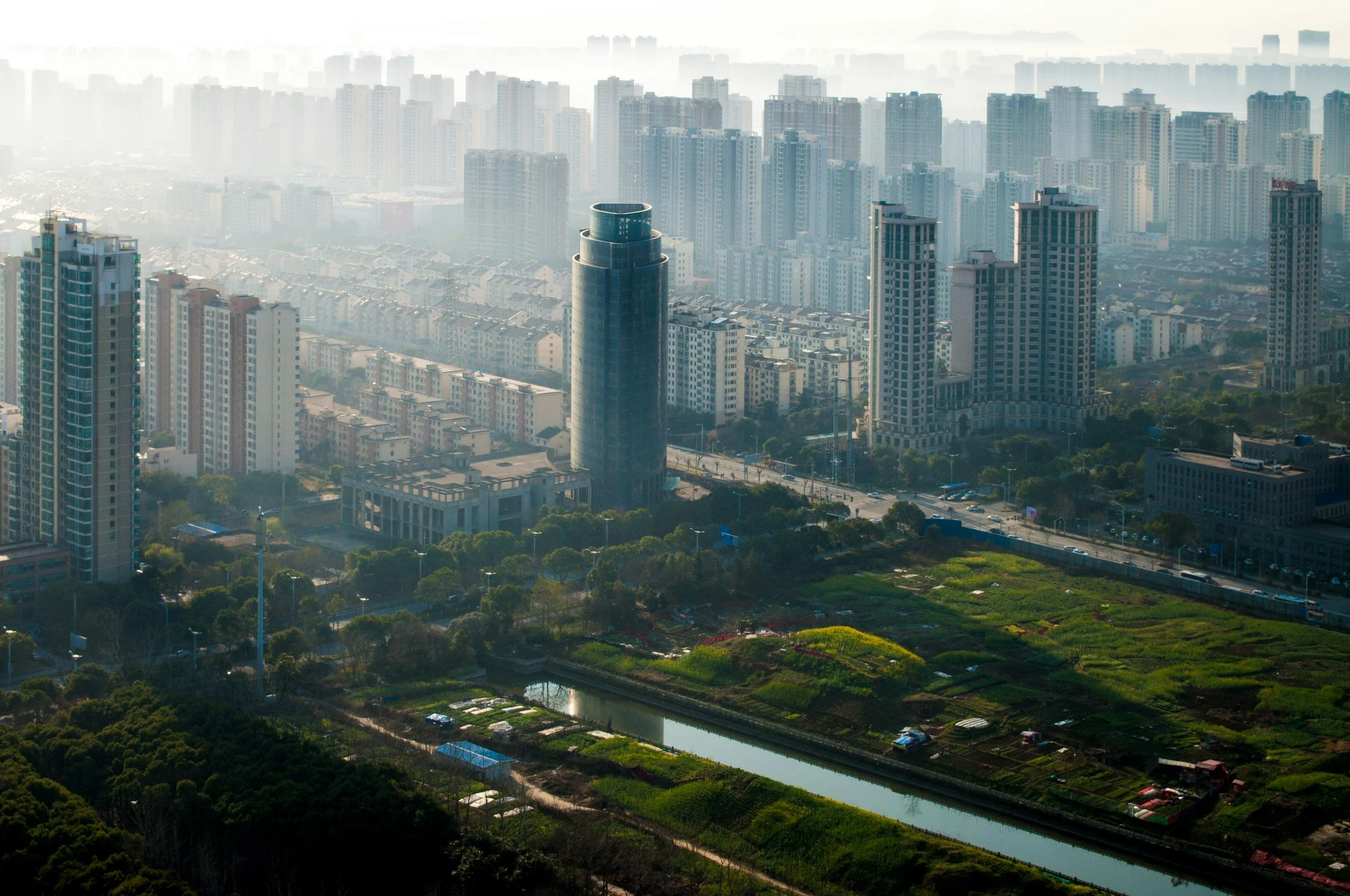 A cityscape showing numerous high-rise buildings with trees, green areas, and a water canal in the foreground, under a hazy sky.