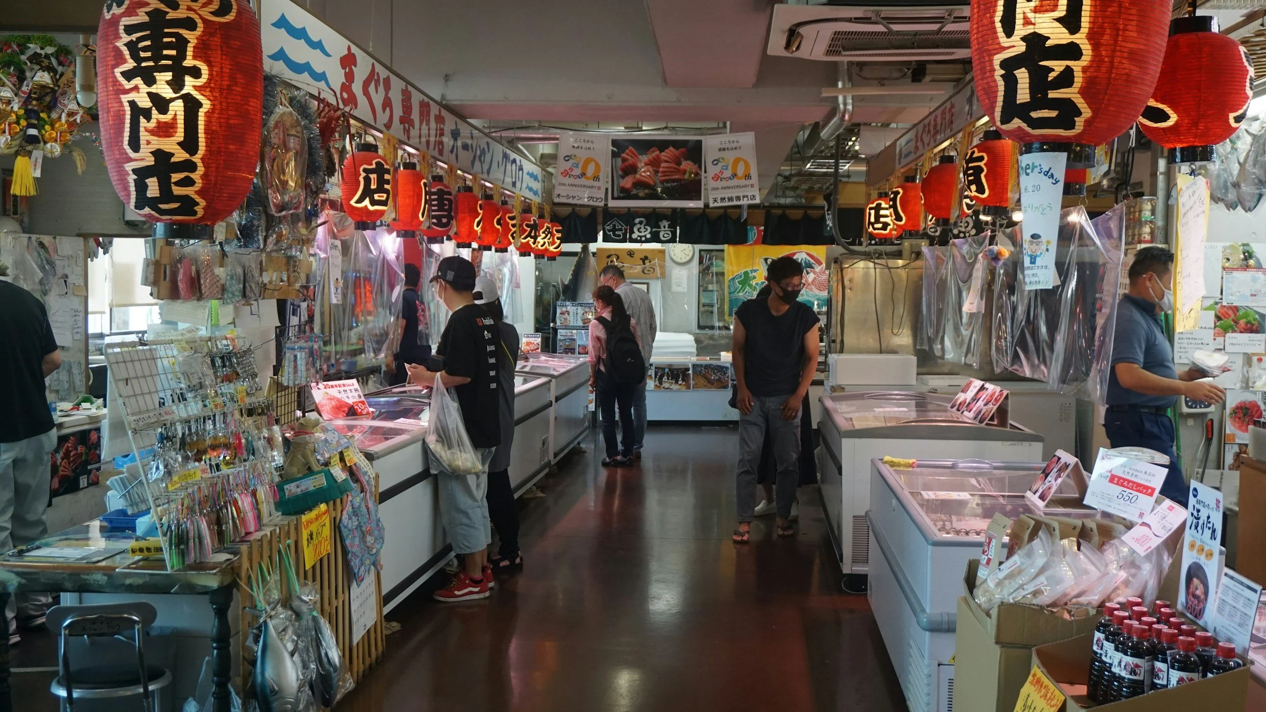 Inside a busy Japanese market with red lanterns hanging from the ceiling, shoppers browsing seafood and various goods at stalls, some wearing masks.