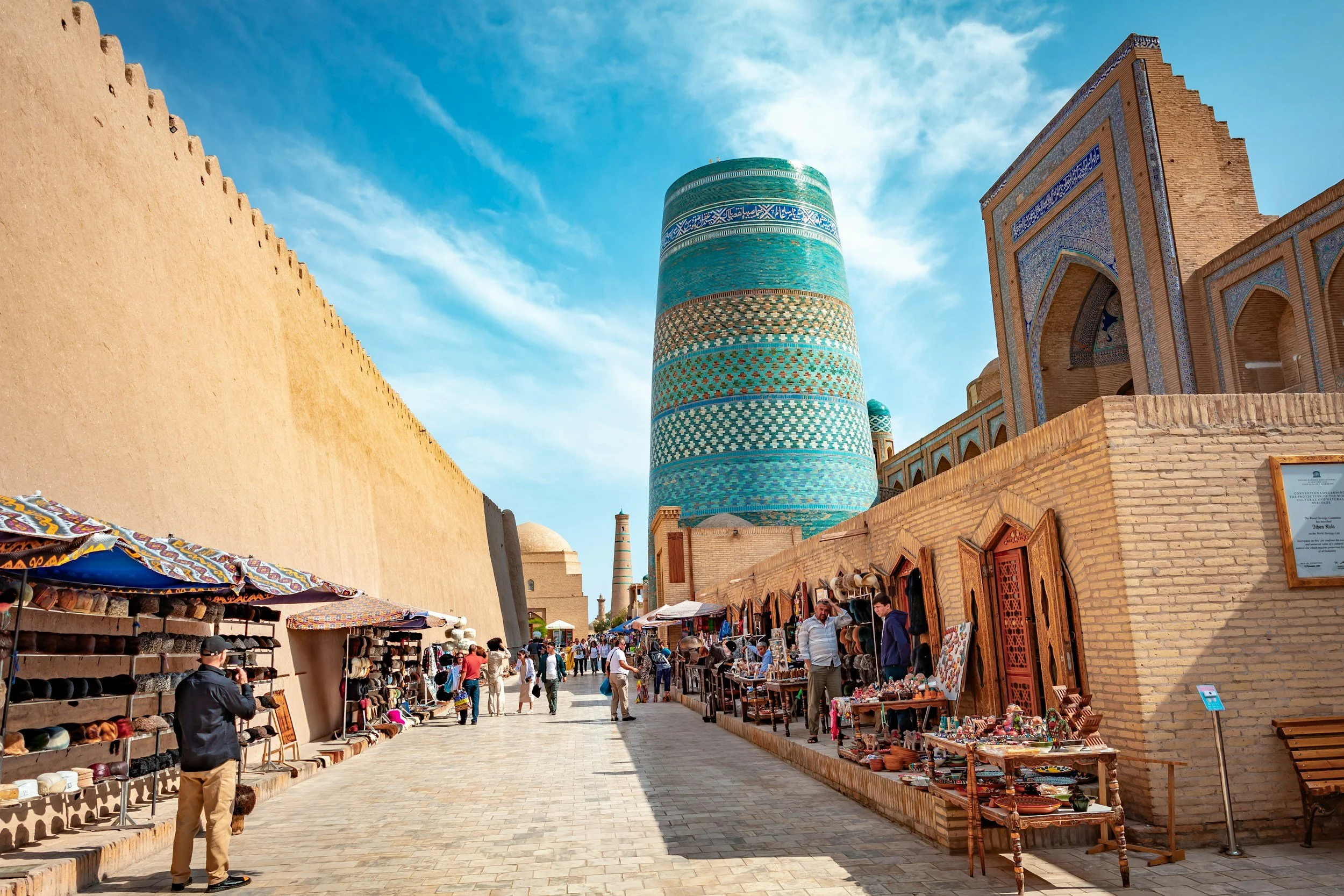 Market street with vendors selling hats and souvenirs, historic architecture, and a tall colorful turquoise minaret in the background, clear blue sky.