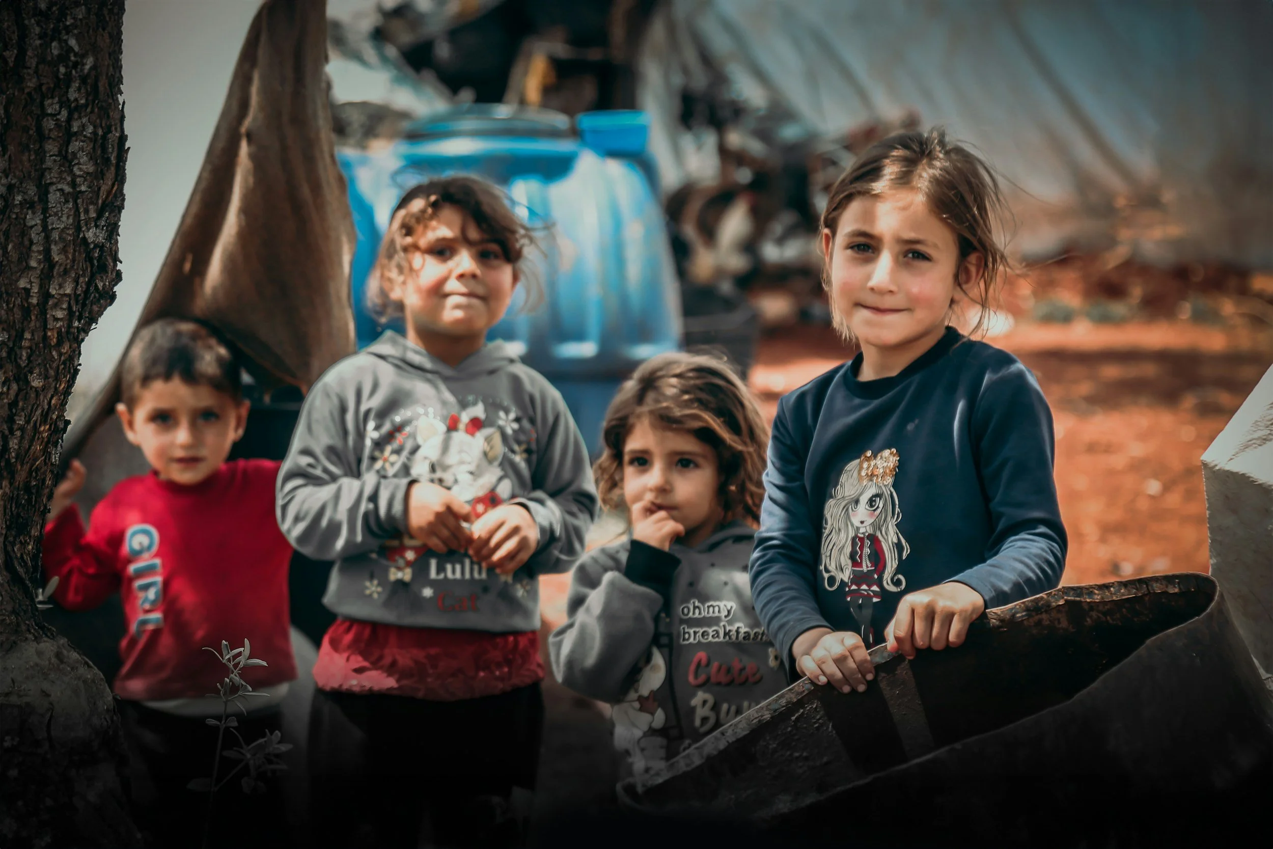 Four children, three girls and one boy, standing outdoors near a tree, with a blurry background of outdoor items and a tent-like structure.