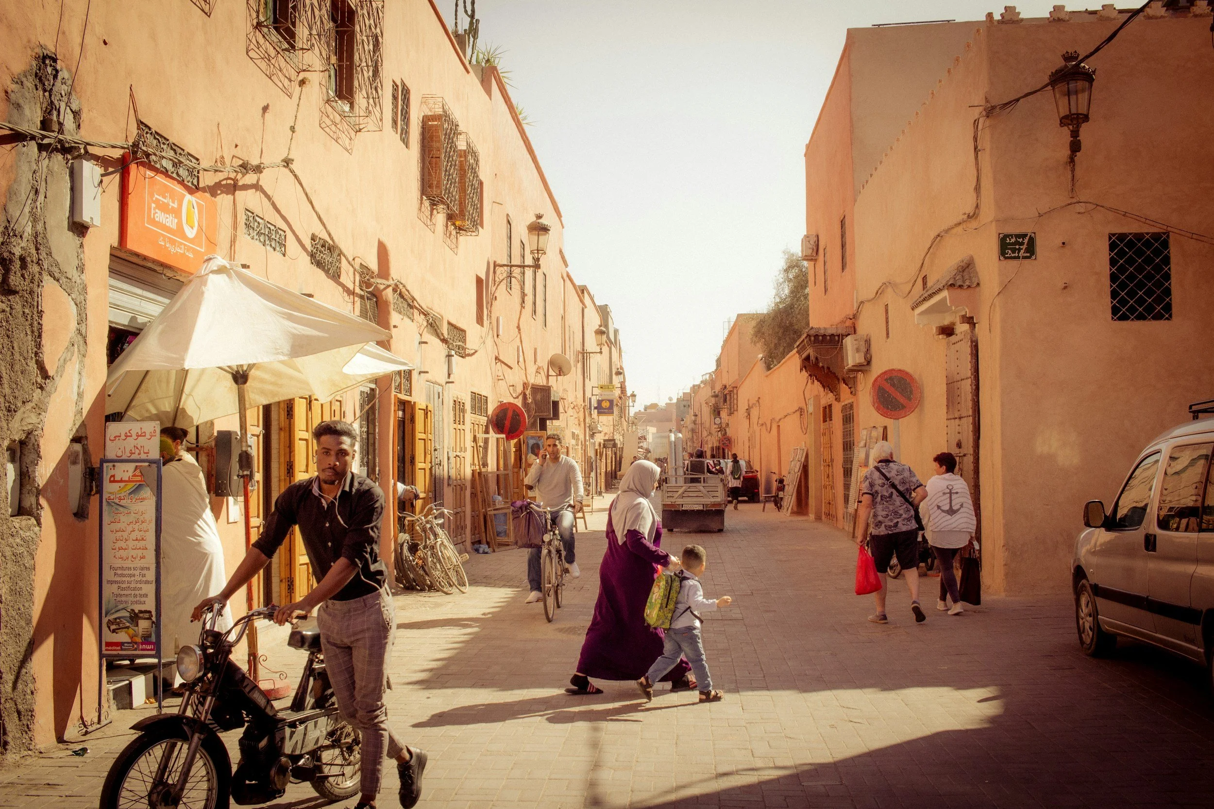 A sunlit street scene with peach-colored buildings, several pedestrians, cyclists, and a man on a motorbike. People are walking, some with bags, and there are bicycles parked along the street. The architecture has small windows with metal grilles and air conditioning units.