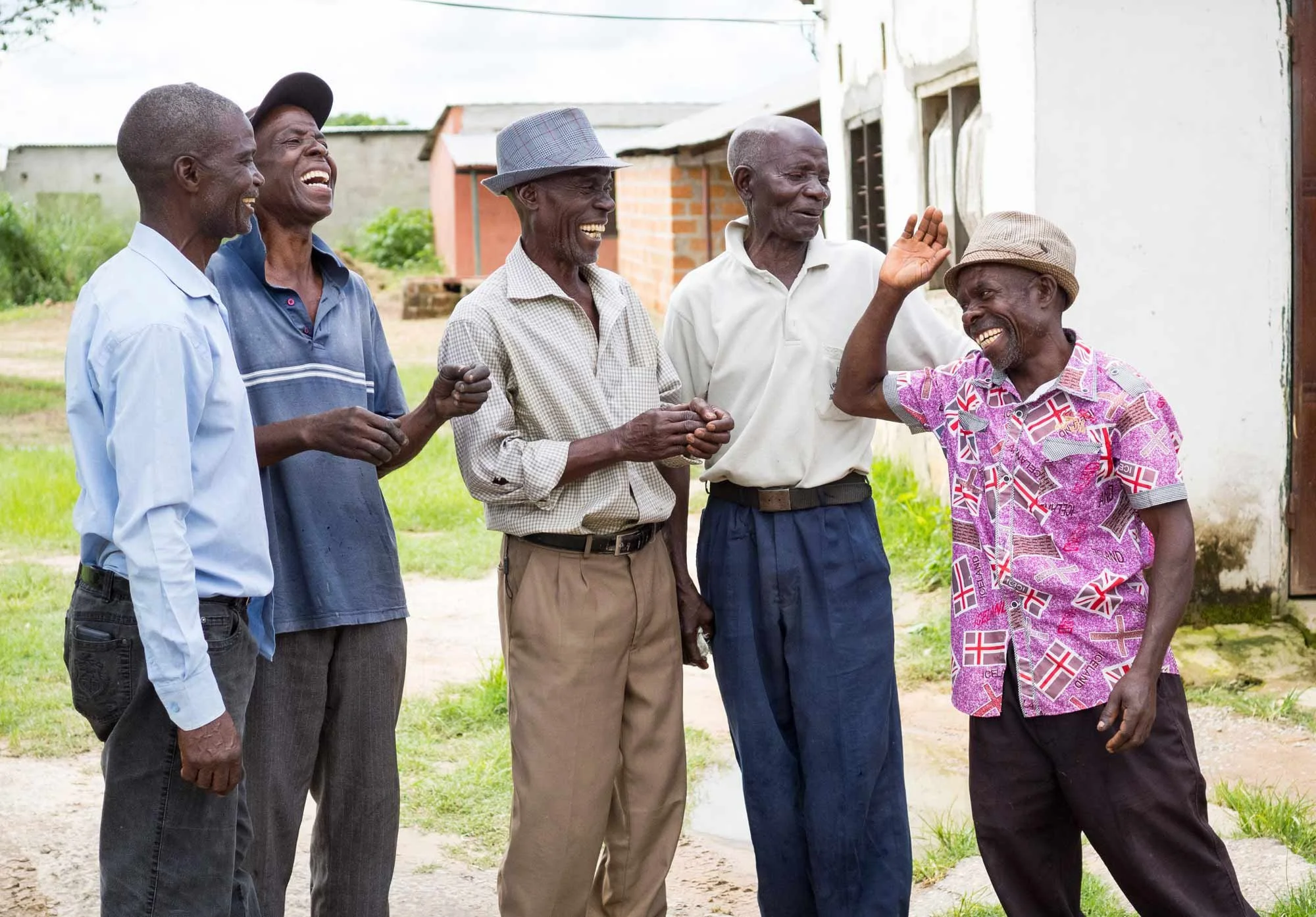 A group of six elderly men talking and laughing outside near a white building, with some small houses in the background.