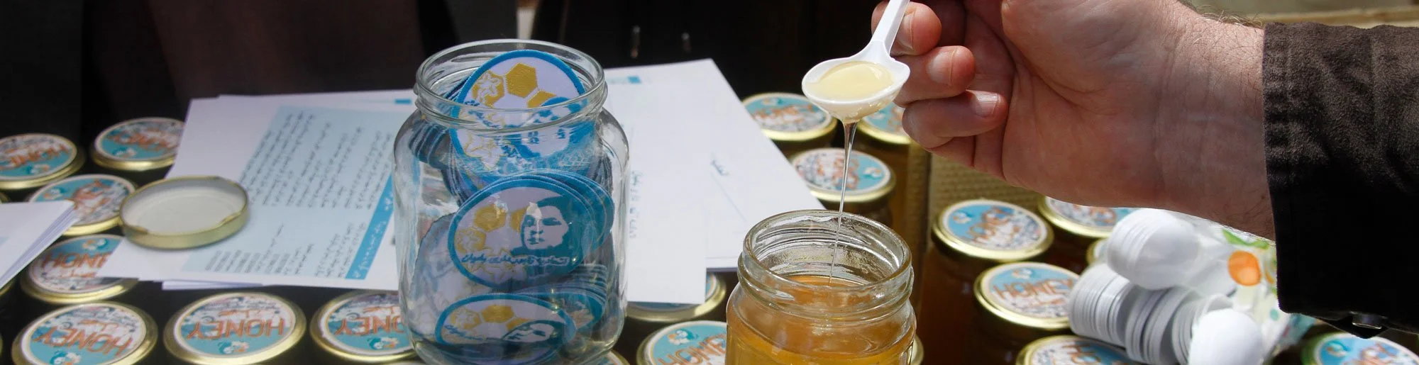 A person is holding a small white spoon with honey over a glass jar of honey, surrounded by jars of honey with labels reading "HONEY" and a document with printed text.