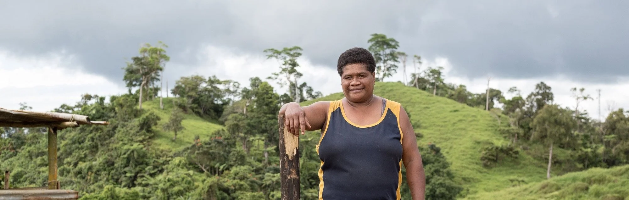 A woman standing outdoors in front of green hills and trees, wearing a black sleeveless top with yellow trim, smiling at the camera.