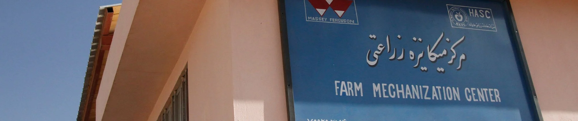 Blue signboard with white text reading 'Farm Mechanization Center' in English and corresponding text in another language, on a light pink building under a clear blue sky.
