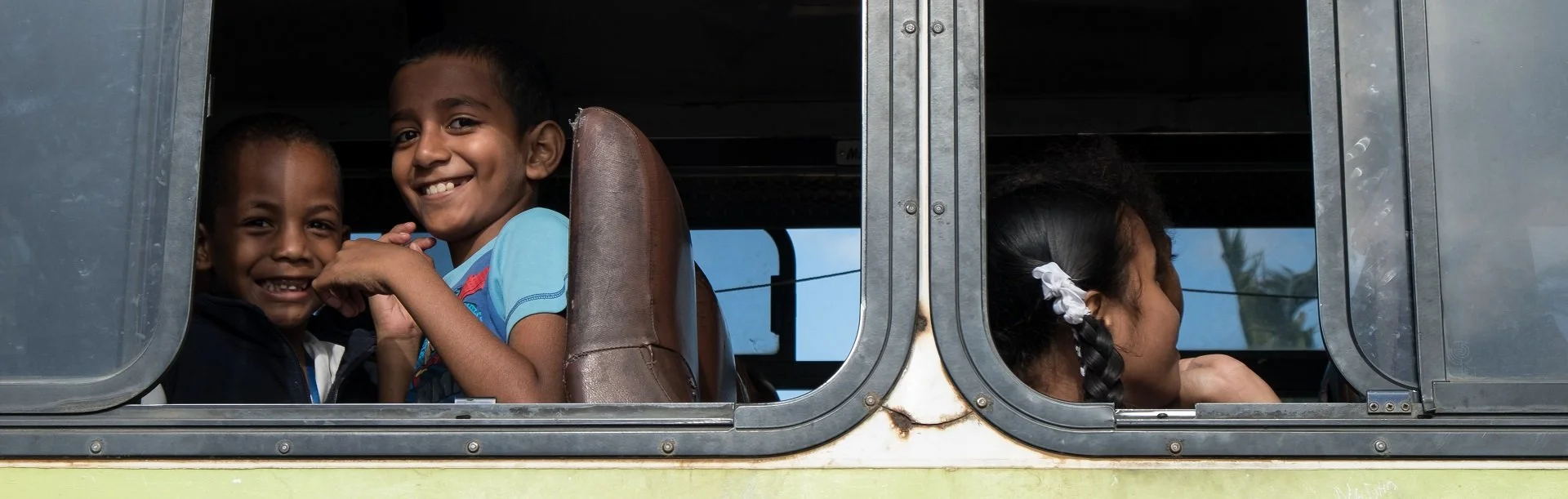 Three children inside a vintage bus, two boys smiling and a girl with her head resting on her hand, looking out the window.