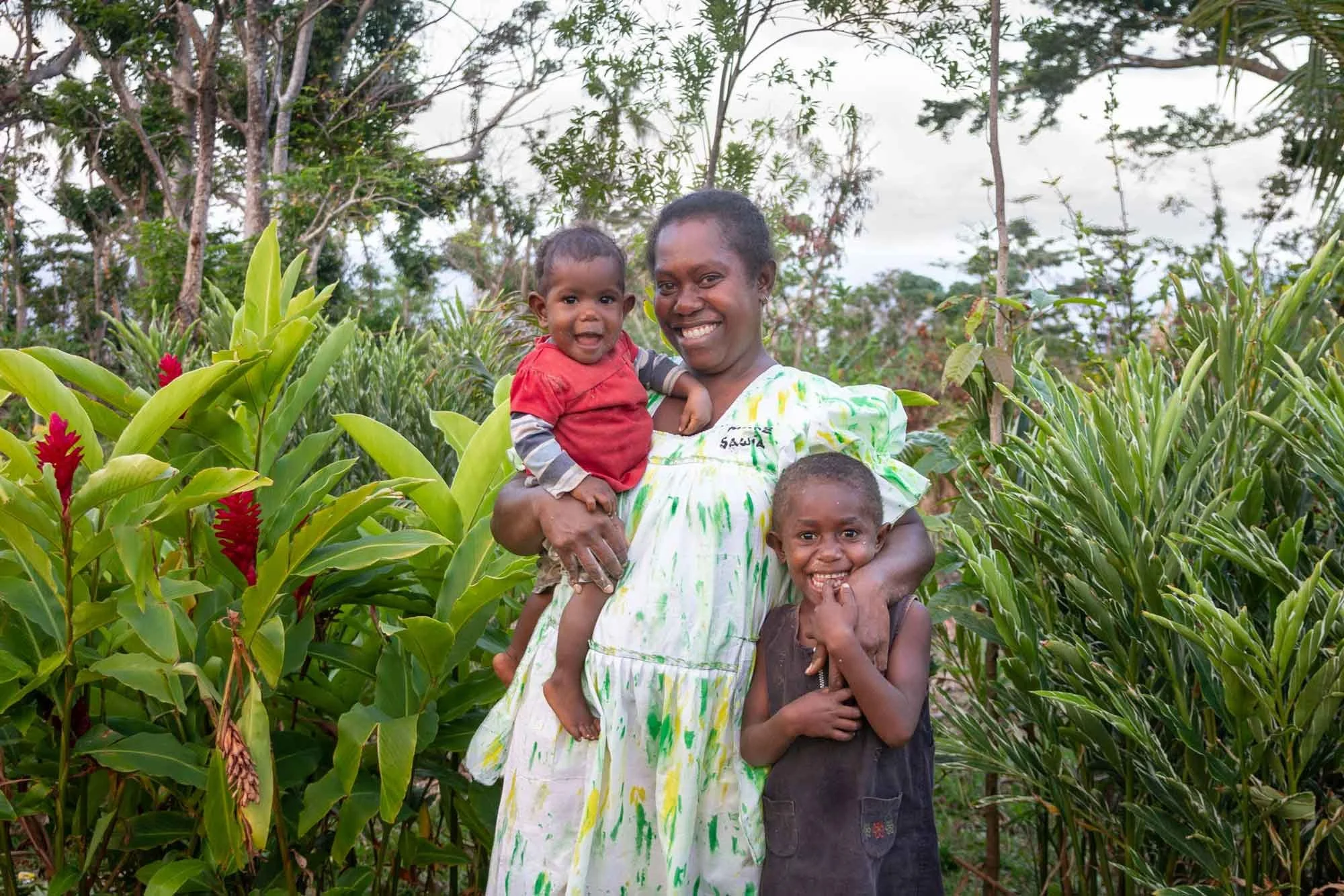 A smiling woman holding two young children in a lush green garden with tall plants and trees.