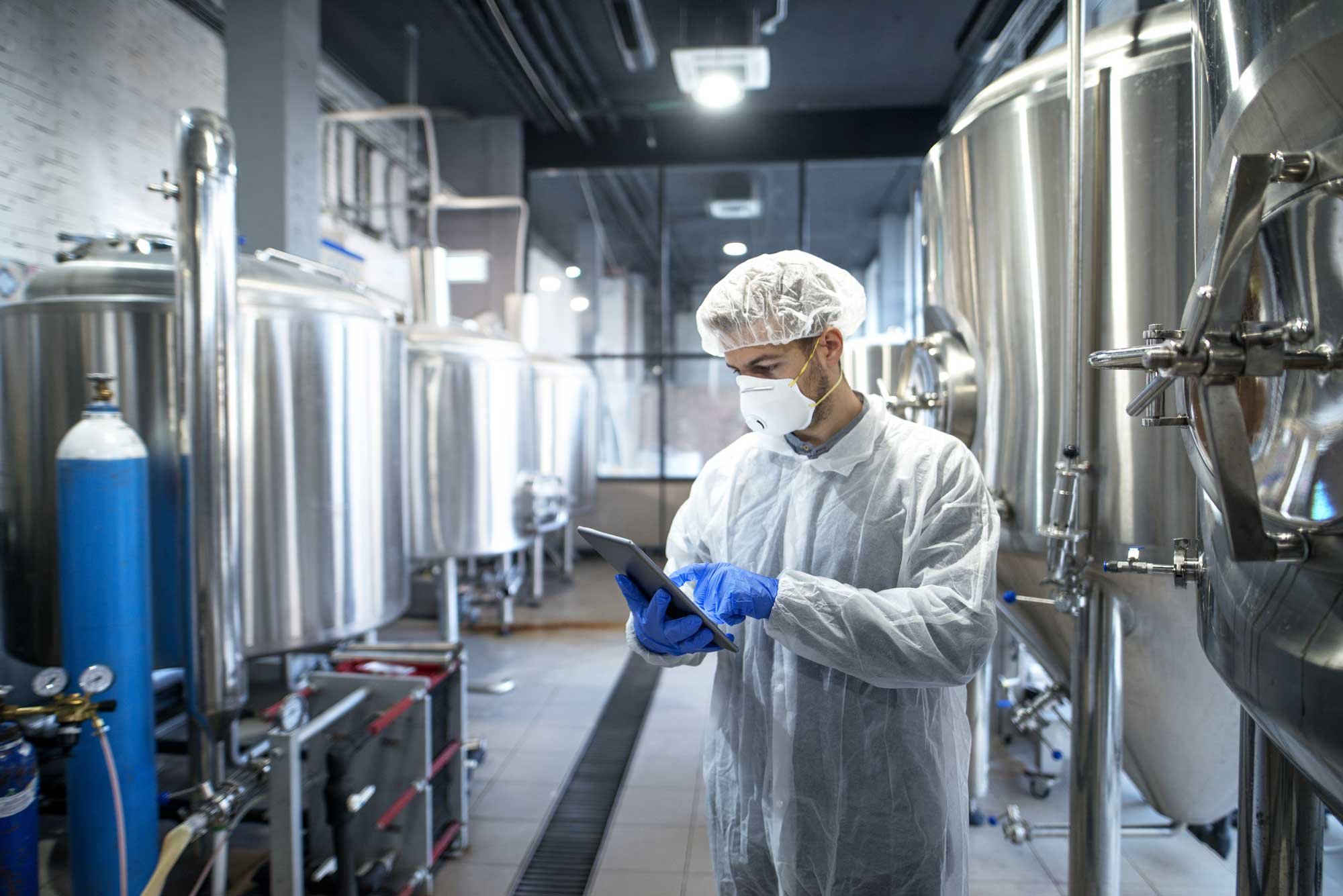 A worker in protective gear, including a mask, hairnet, gloves, and lab coat, stands in a brewery or industrial kitchen, looking at a tablet with large stainless steel tanks in the background.