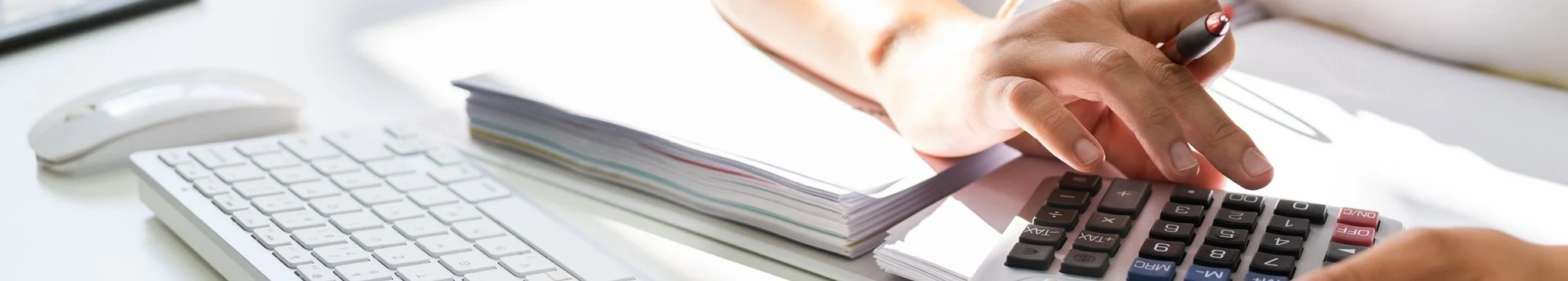 Close-up of a person’s hand pressing a calculator key at a desk with a white keyboard, a mouse, and a stack of papers.