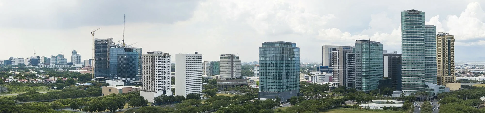 City skyline with tall buildings and green trees in the foreground under partly cloudy sky.