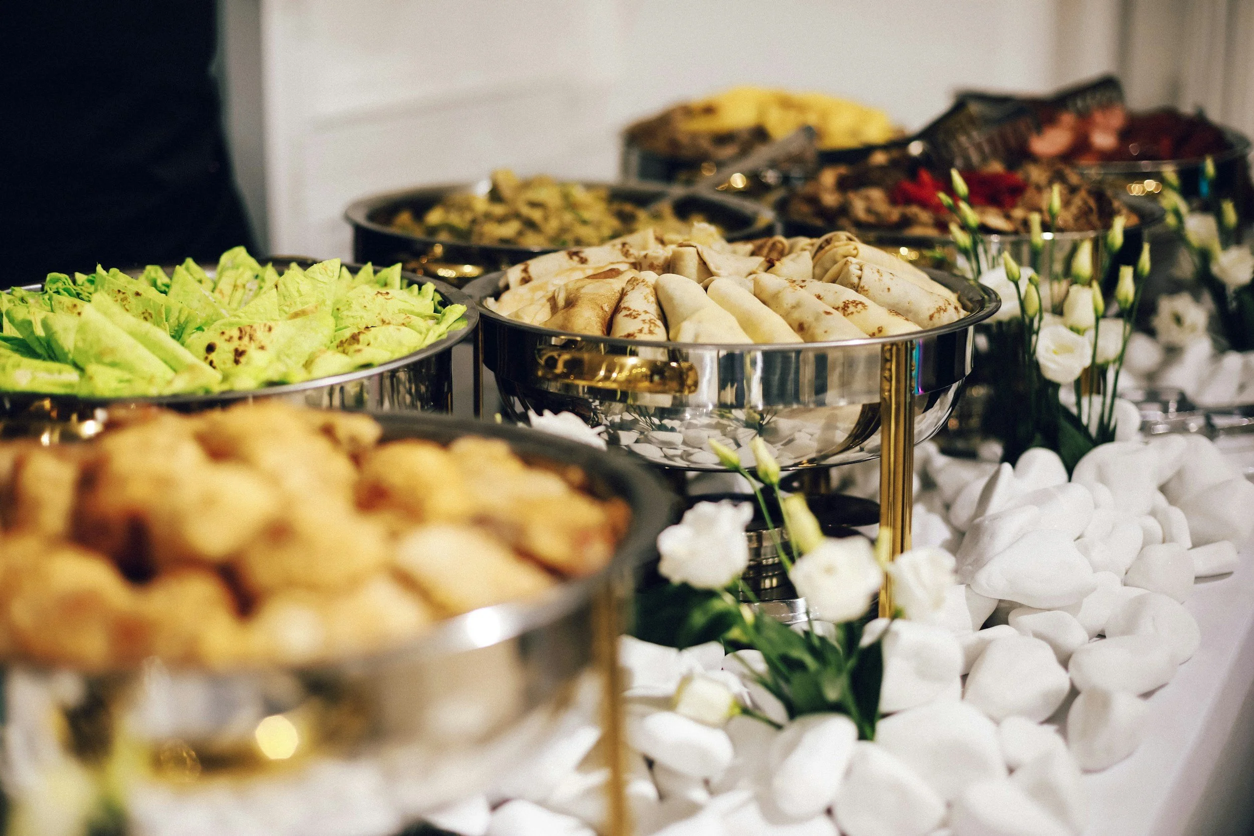 A buffet table with various dishes including spring rolls, lettuce wraps, and other finger foods, decorated with white flowers and stones.