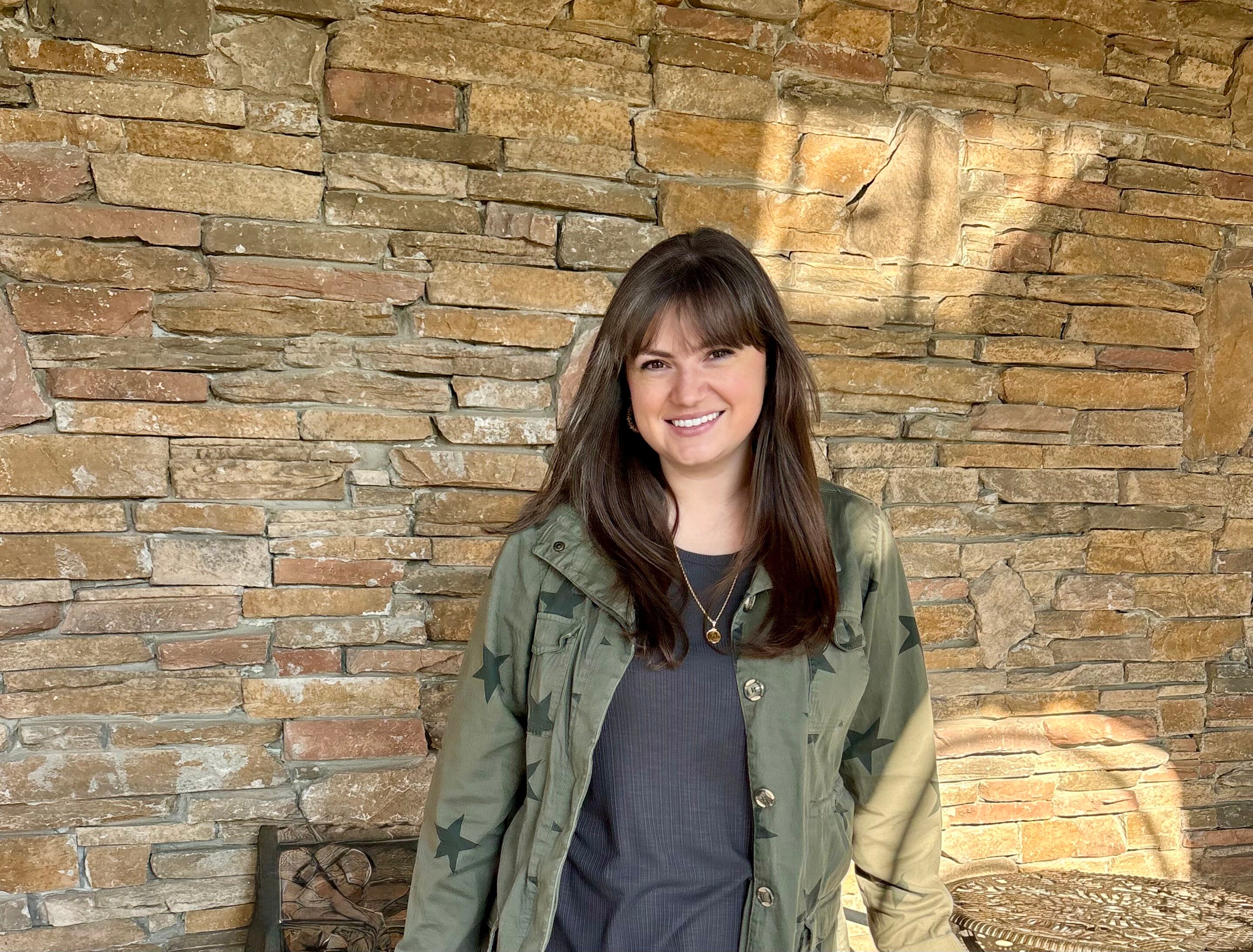A young woman with long brown hair, wearing a green jacket with star patterns, a black top, and a gold necklace, smiling in front of a stone wall.