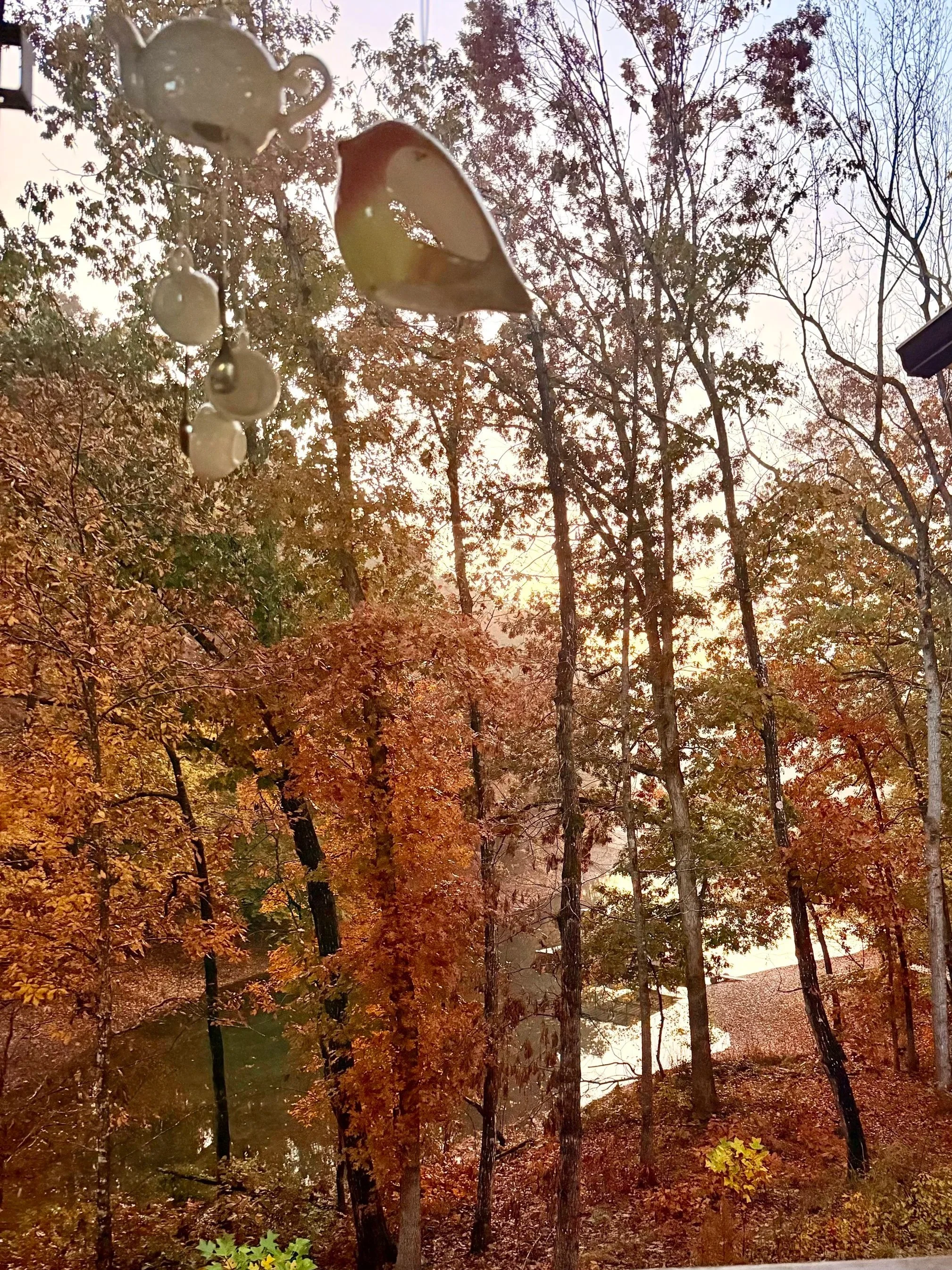 A window with a hanging decoration consisting of a small teapot, a bird shape, and three white beads, with a view of a wooded area with fall-colored trees and a body of water in the background.