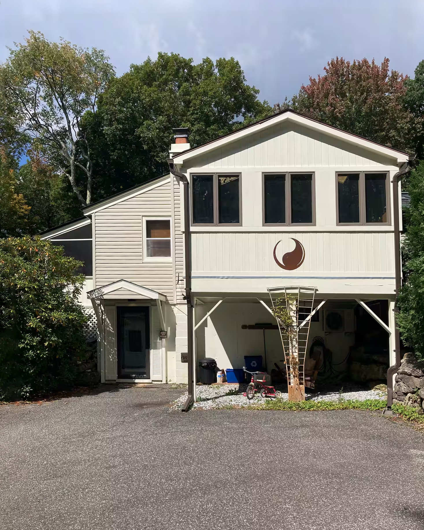 Front view of a two-story house with light-colored siding, large upper windows, a small porch, and a driveway, surrounded by trees.