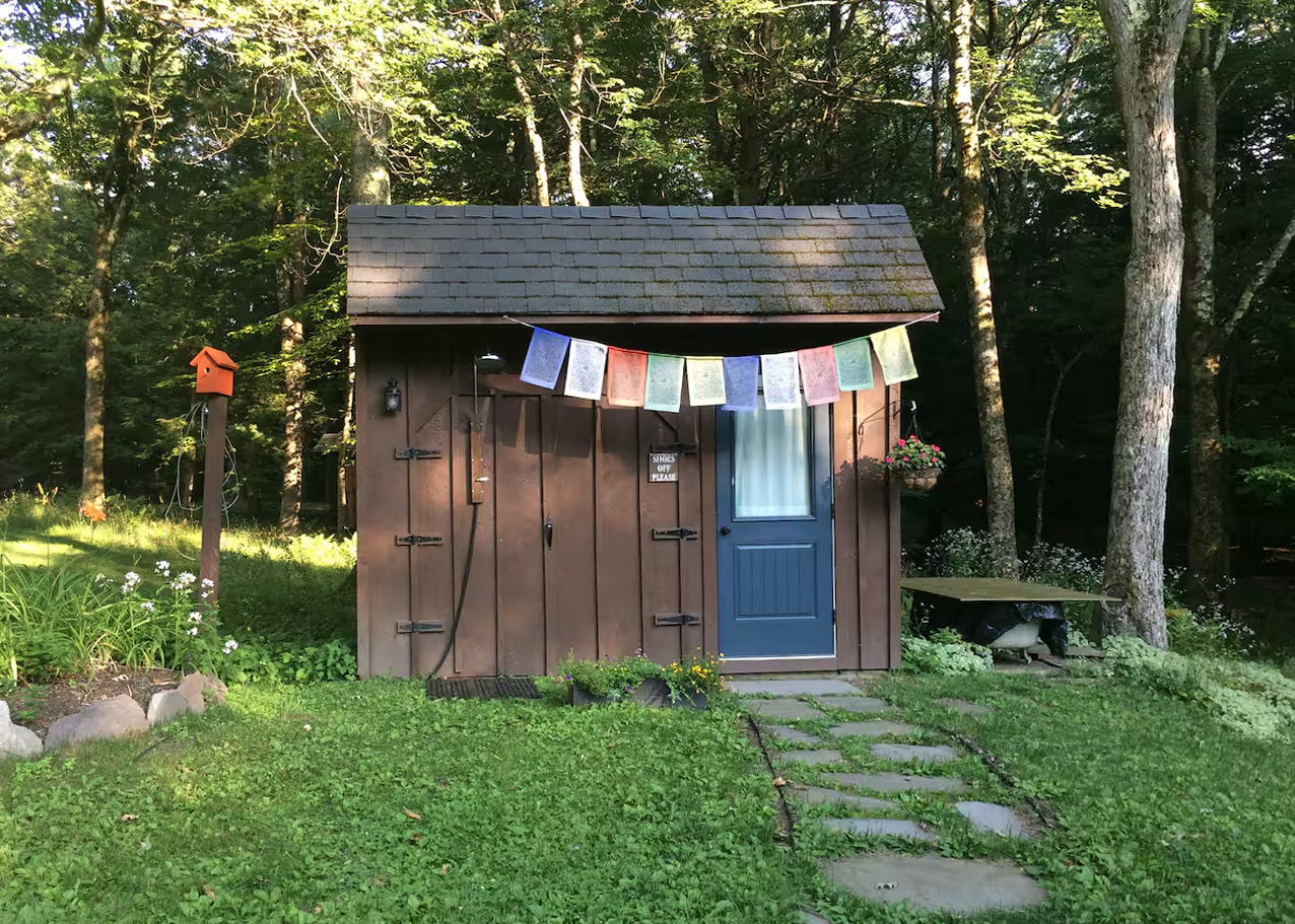 A small wooden shed with a blue door and blue window in a green garden, surrounded by trees. There are colorful prayer flags hanging in front of the shed and a birdhouse on a post to the left. A stone pathway leads to the door, and there are flower beds on the left and a wooden bench on the right.