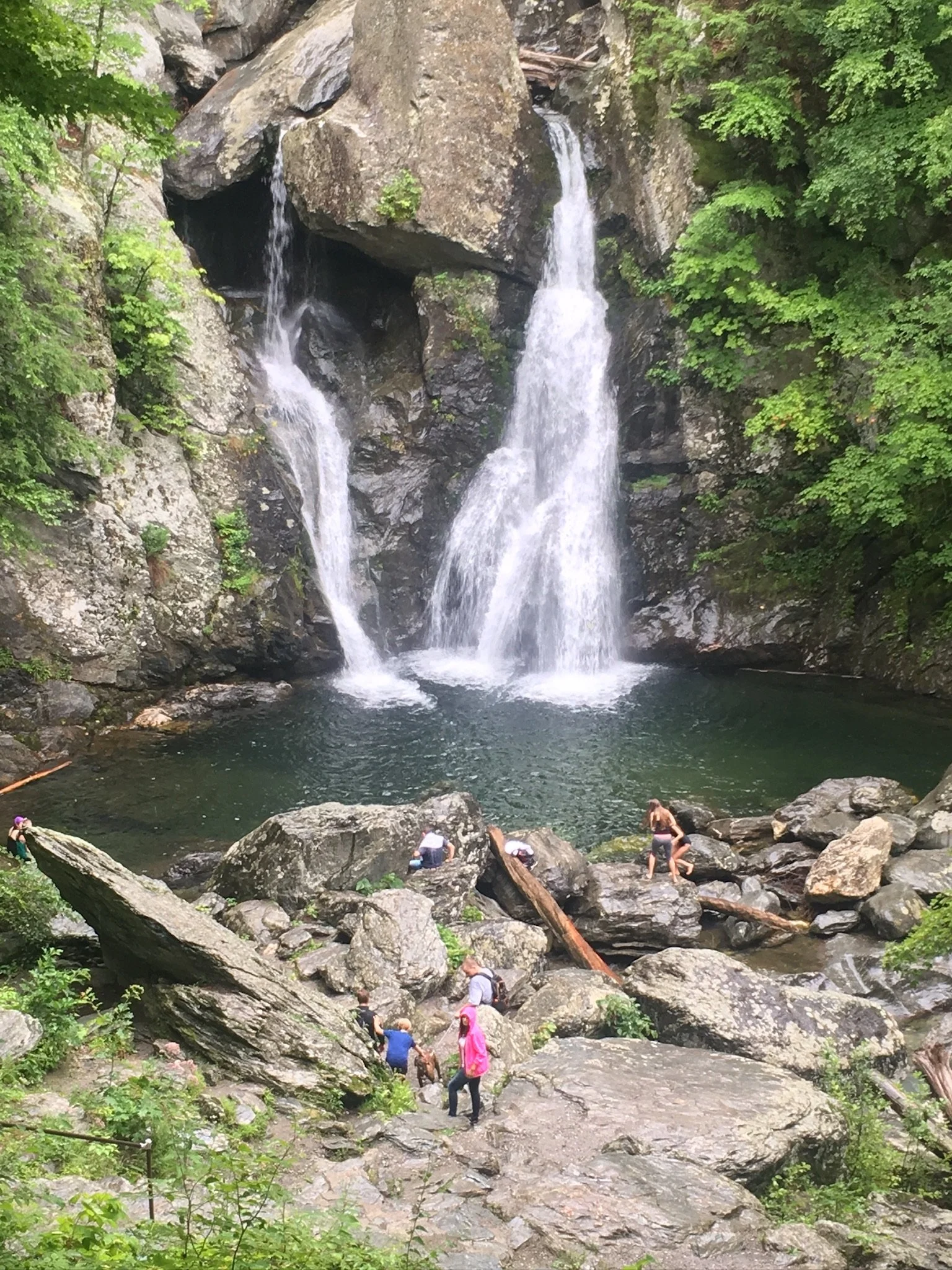 People exploring rocks and hiking near a double waterfall in a lush forest