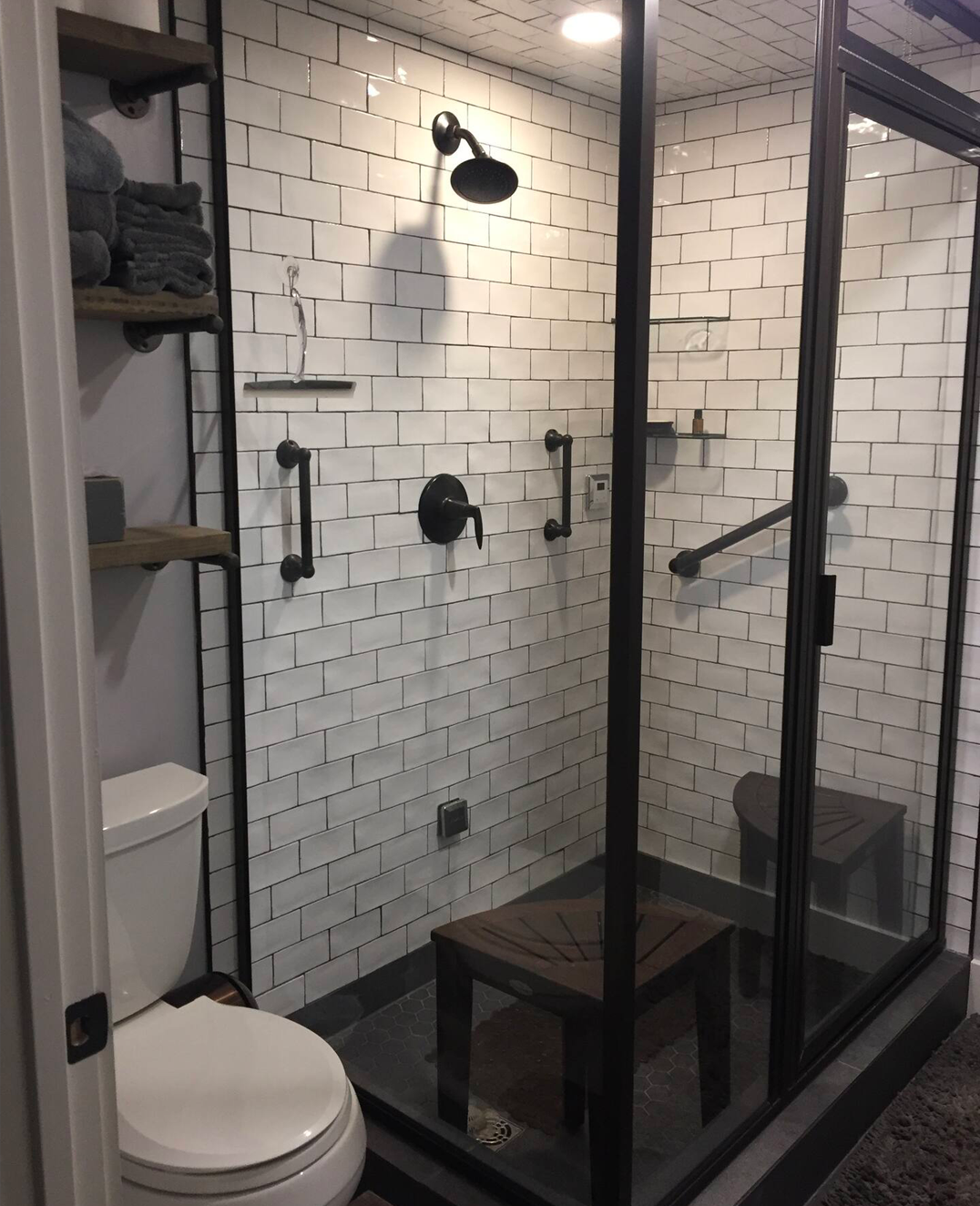 Modern walk-in shower with black fixtures, glass door, white subway tile walls, wooden benches, and shelves with towels in a bathroom.