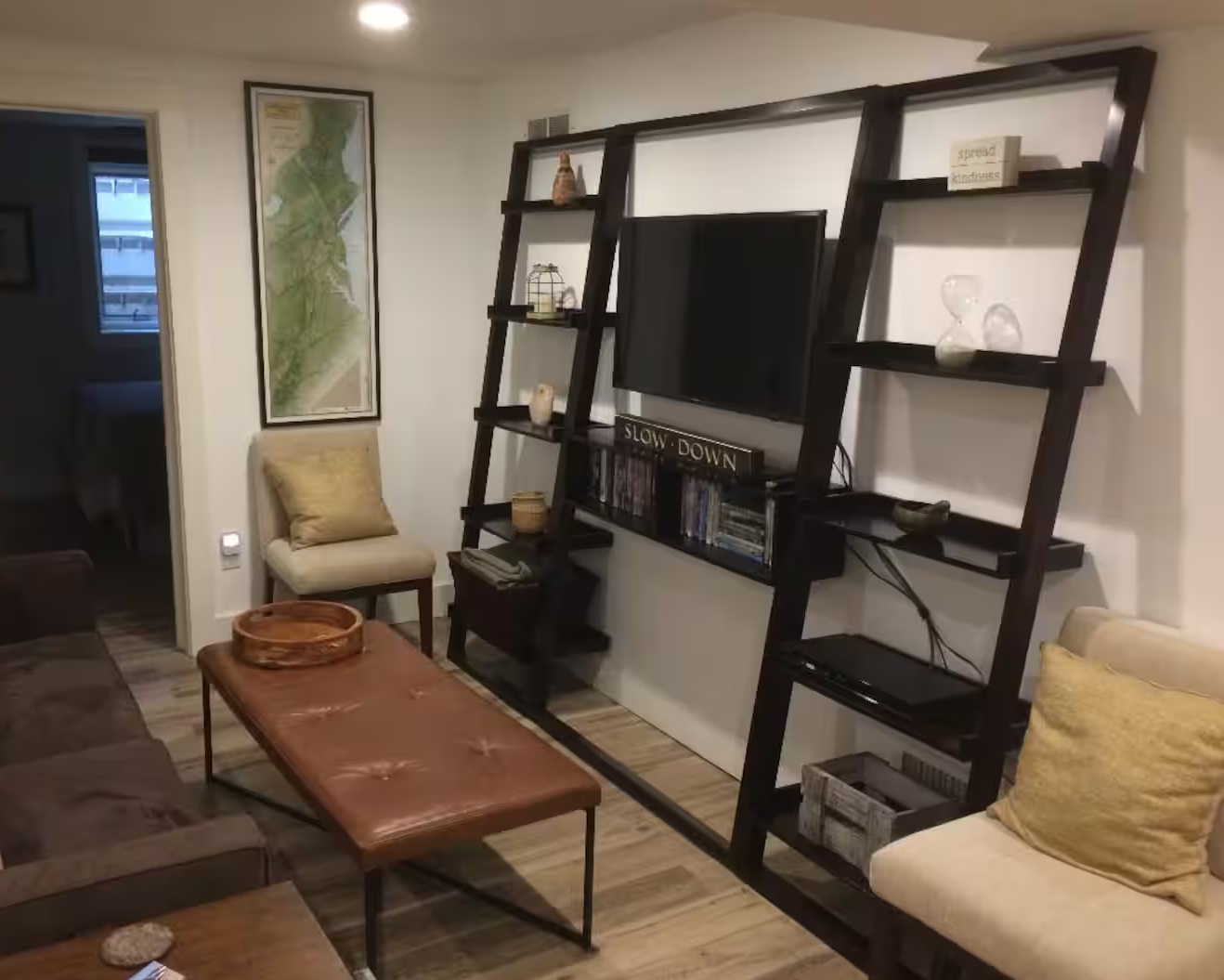 Living room with a wooden ladder-style bookshelf, a flat-screen TV, a beige armchair with a pillow, a brown sofa, a wooden coffee table with a tray, a framed map on the wall, and decorative items on shelves.