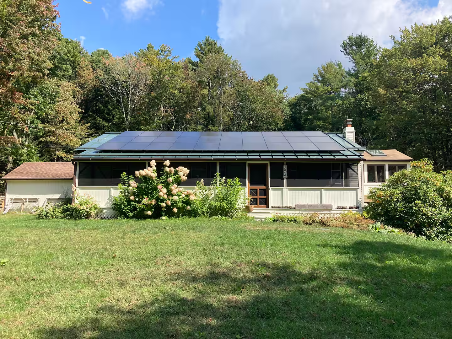 A house with solar panels on the roof, surrounded by greenery and flowering bushes in a lawn, with trees in the background under partly cloudy sky.