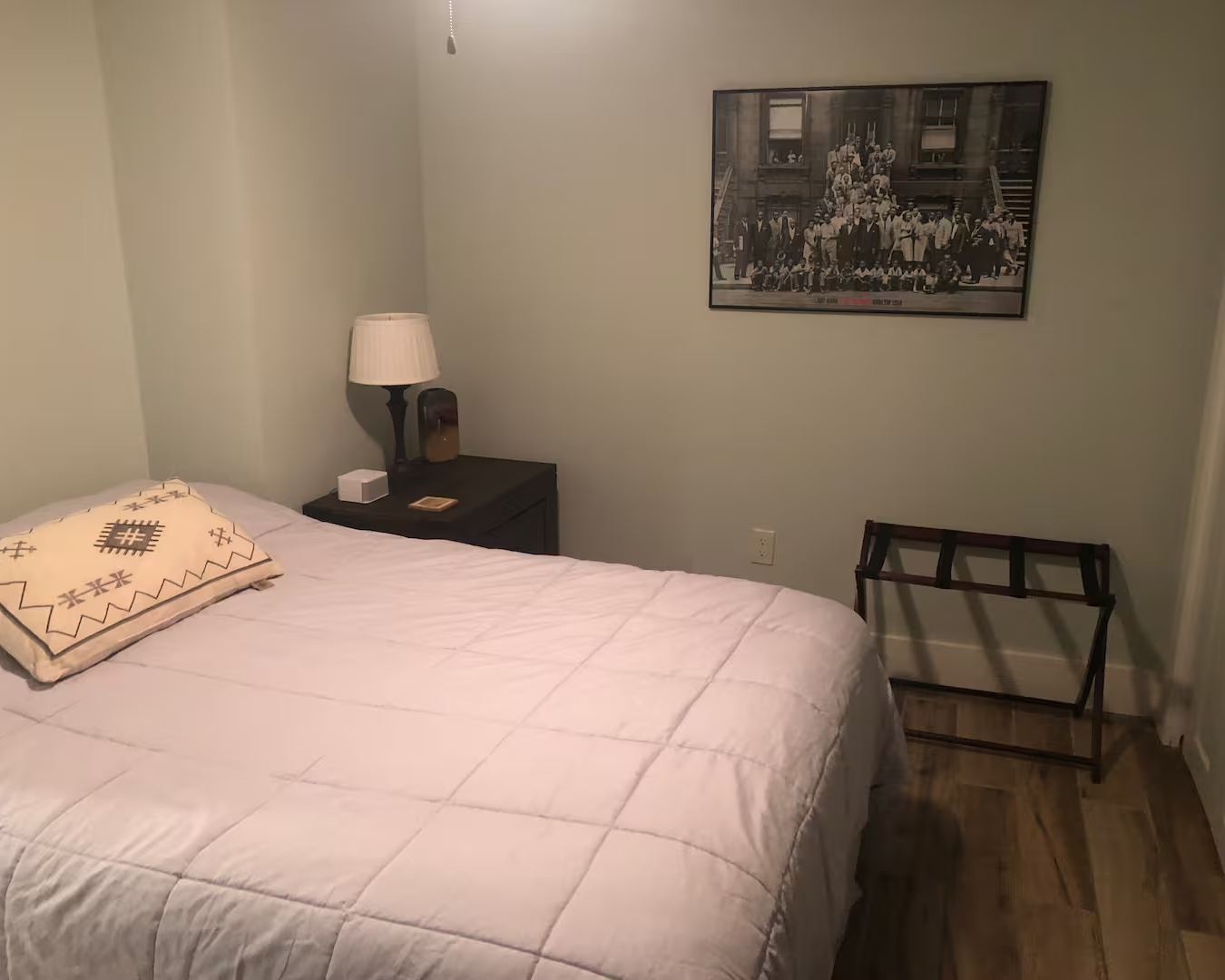 A bedroom with light green walls, a bed with white bedding and a decorative pillow, a dark wood nightstand with a lamp, a framed black and white group photo on the wall, and a luggage rack in the corner.
