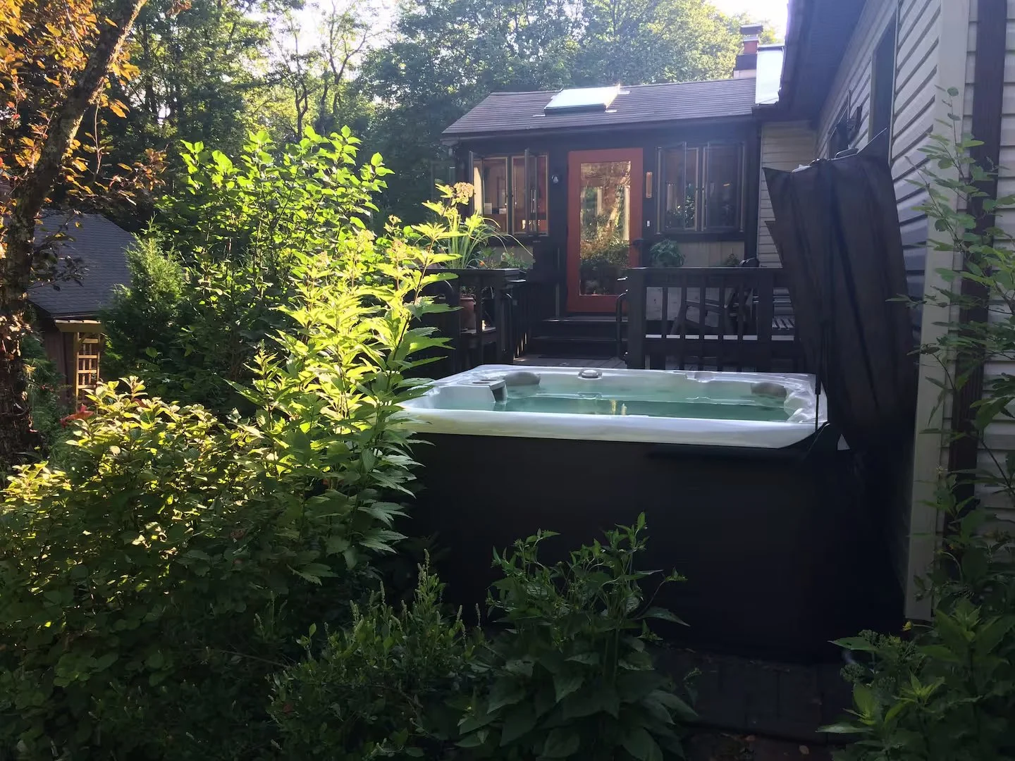 A backyard with a hot tub on a deck, surrounded by greenery, and a house with a large window and a door leading outside.
