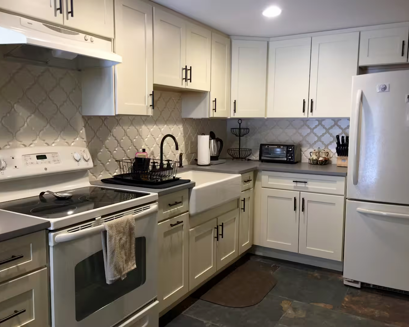 Kitchen with white cabinets, a white stove with a towel hanging on the handle, white farmhouse sink, black faucet, beige tiled backsplash, small countertop appliances, paper towel roll, and a refrigerator.