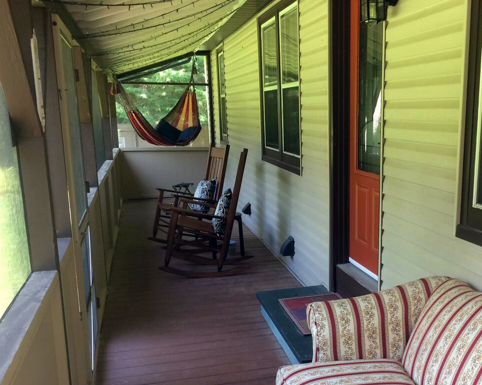 A porch with wooden flooring, two rocking chairs with patterned cushions, a red door, a floral-patterned couch, and a colorful hammock hanging from the ceiling, overlooking trees.