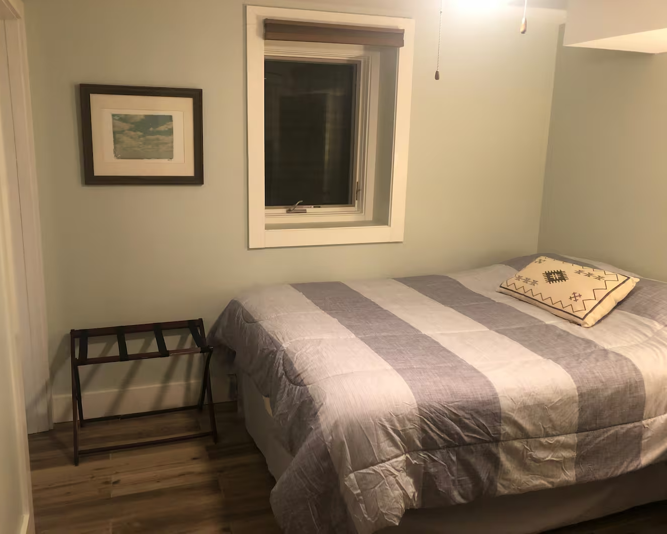 A bedroom with a bed covered in a striped gray and white quilt, a decorative pillow, a window above the bed, a picture on the wall, and a luggage rack beside the bed.