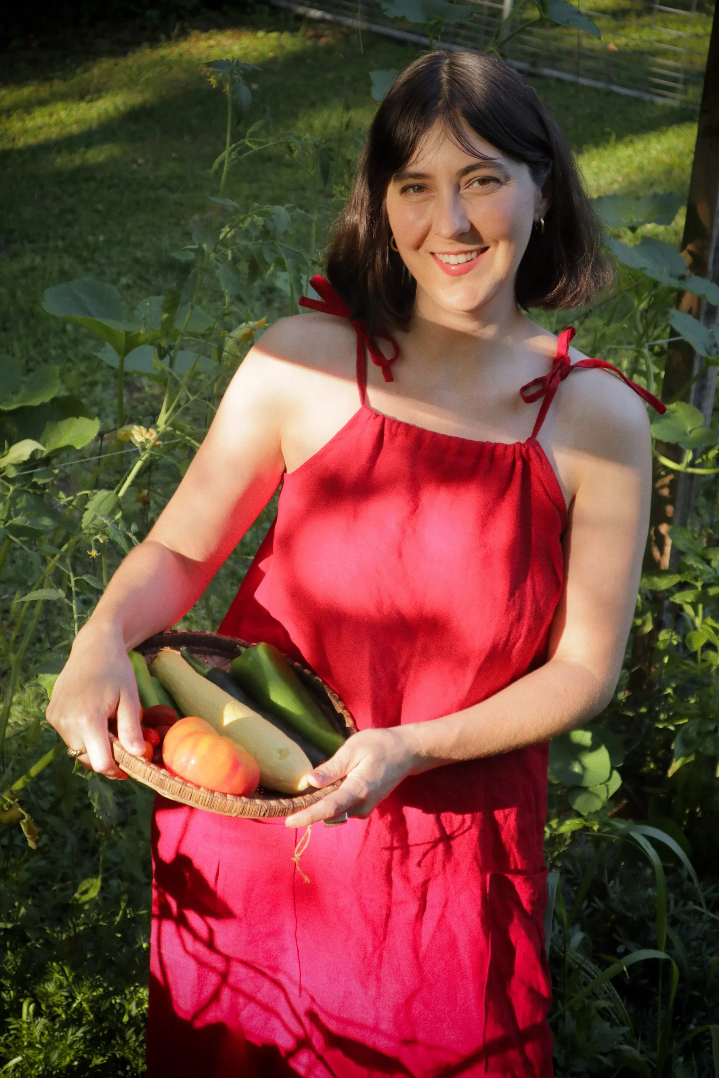 A woman in a red dress holding a basket of fresh vegetables in a garden.