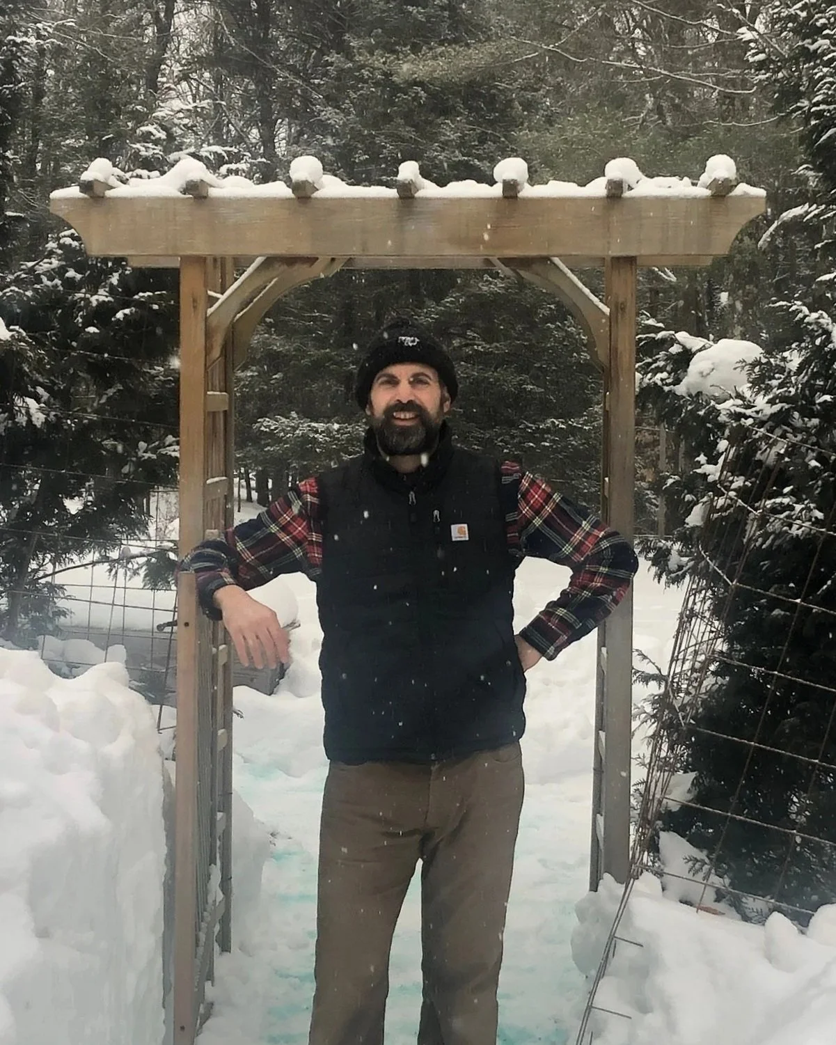 A man with a beard and a black beanie hat standing outdoors in a snowy landscape. He is smiling, wearing a plaid shirt, a black vest, and tan pants, and has his hands on his hips. He stands under a wooden structure with snow on top, surrounded by snow-covered trees and a wire fence.