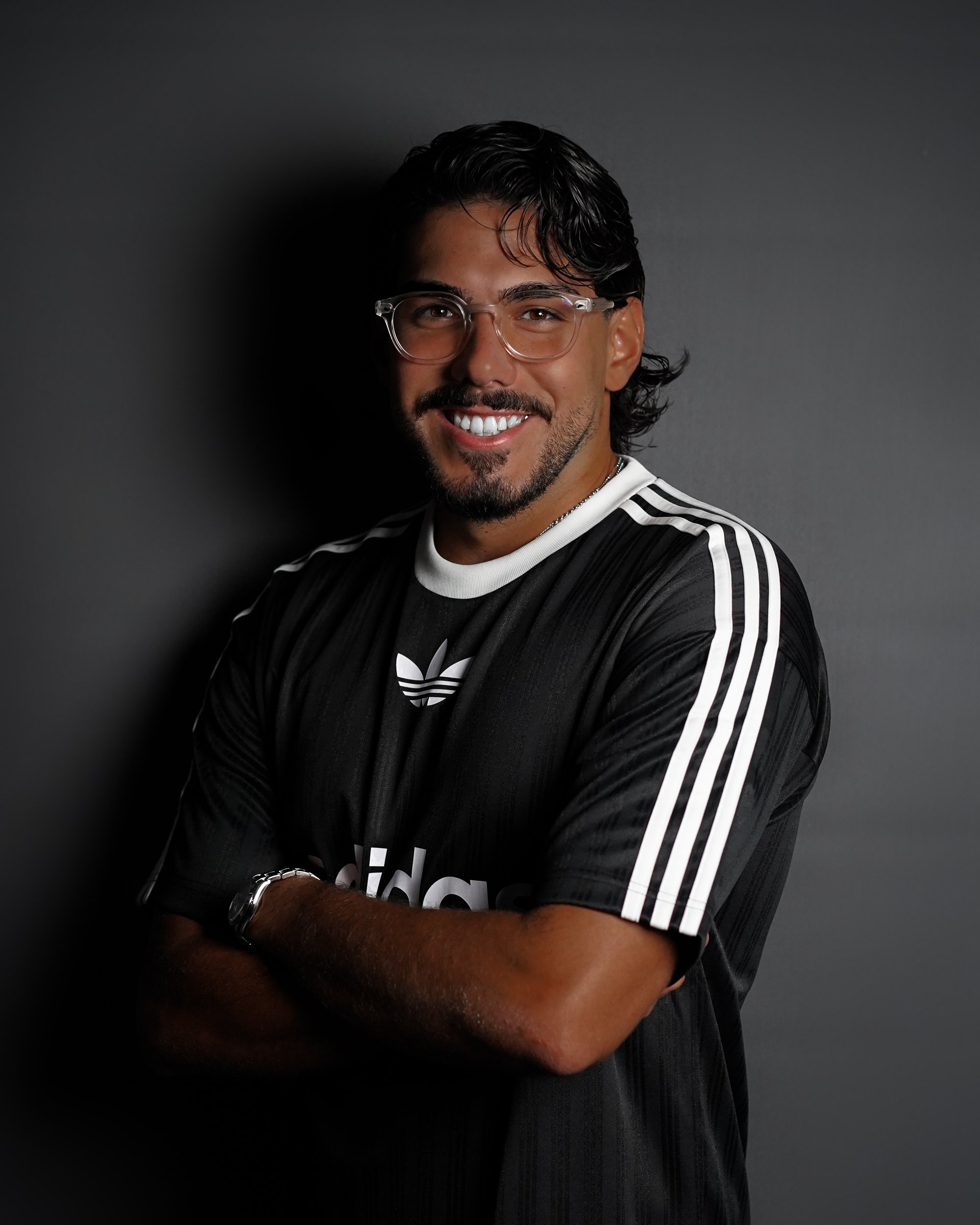 A smiling young man with dark hair, beard, and glasses wearing a black Adidas sports jersey with white stripes on the sleeves, standing against a dark background.