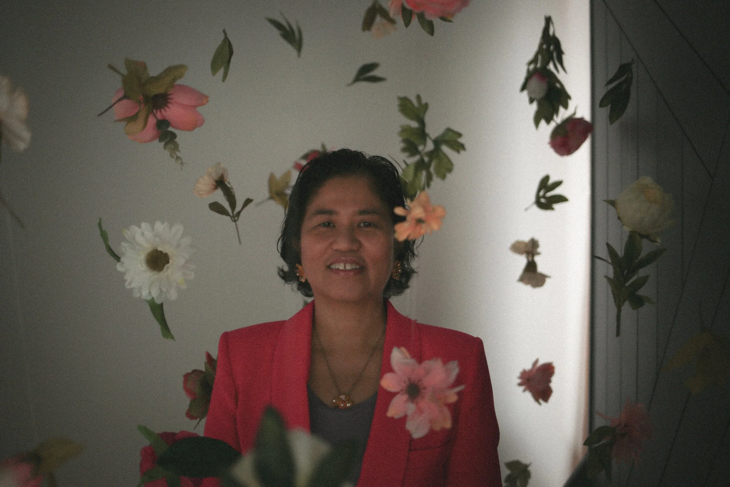 A woman wearing a red blazer smiling at the camera, surrounded by floating pink and white flowers in an indoor setting.