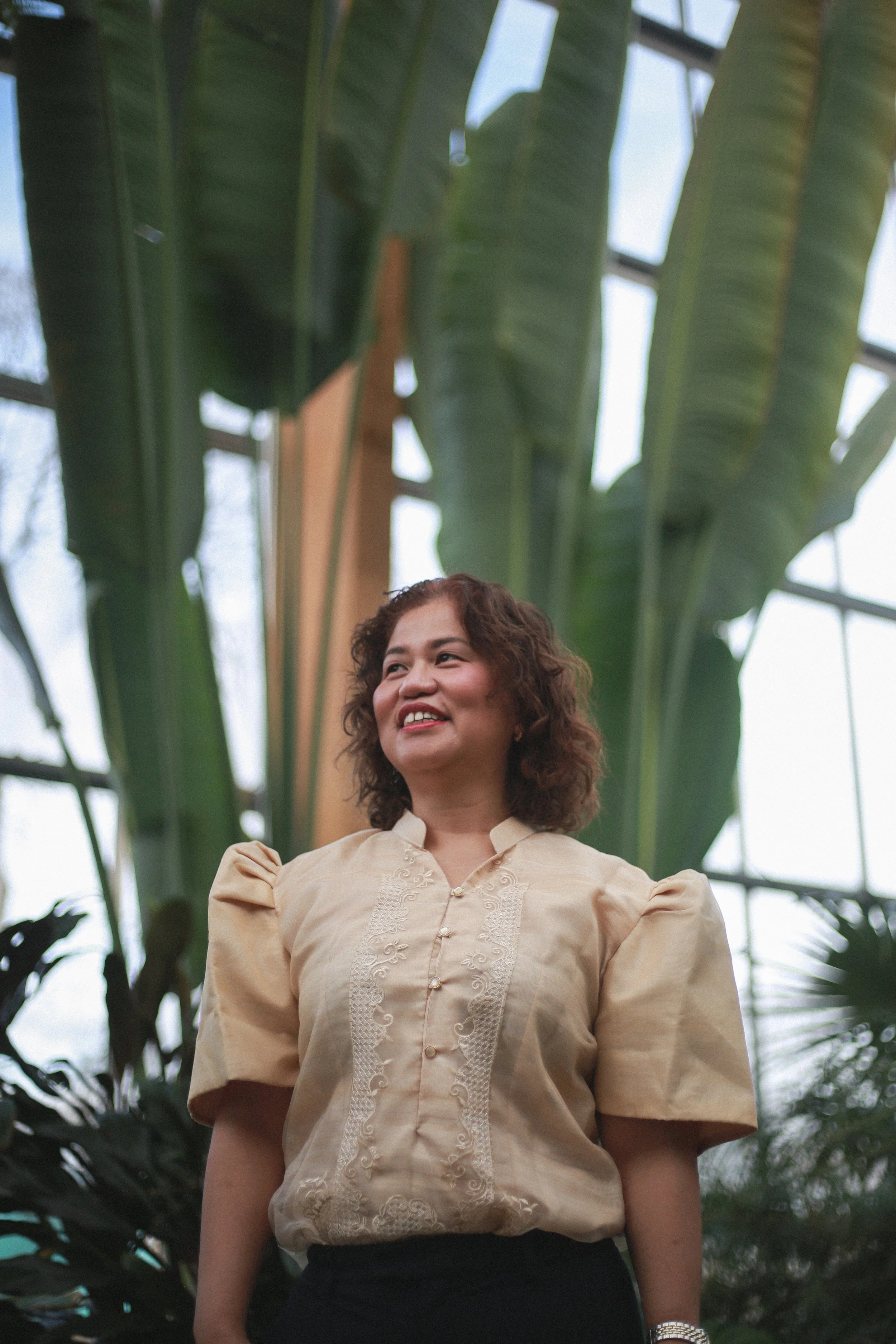 A woman standing inside a greenhouse with large green leaves behind her, smiling and looking upward.