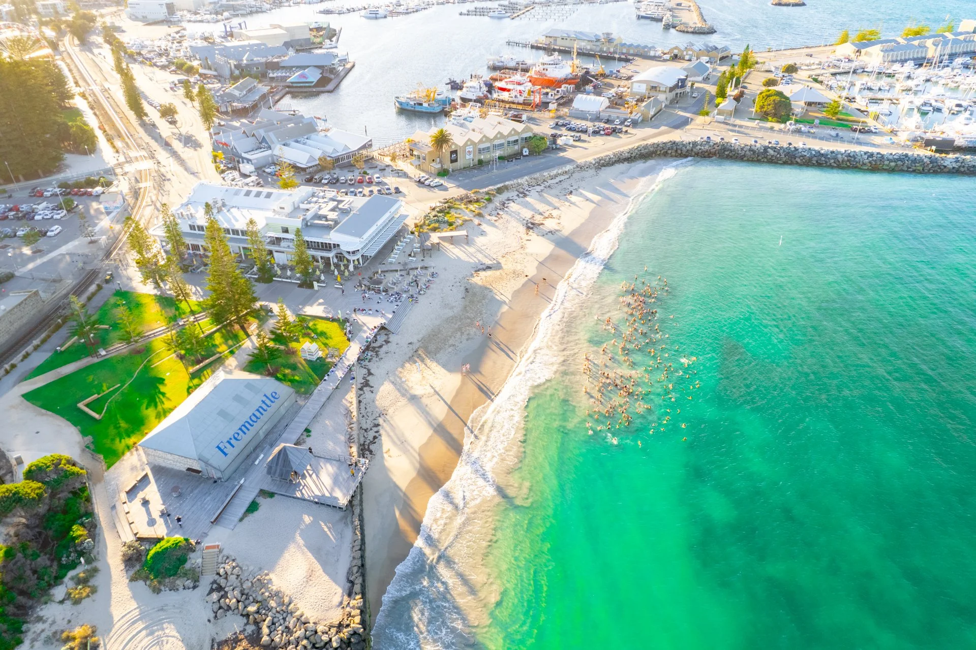 Aerial view of a beach and marina in Fremantle with a greenish ocean, sandy shore, and boats docked at the harbor.