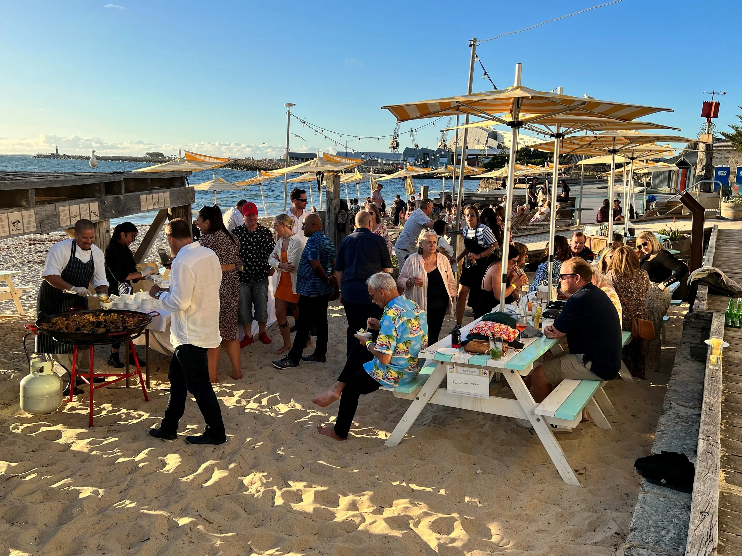 Guests at a function on the sand at Bathers Beach House, with paella being served from the pan