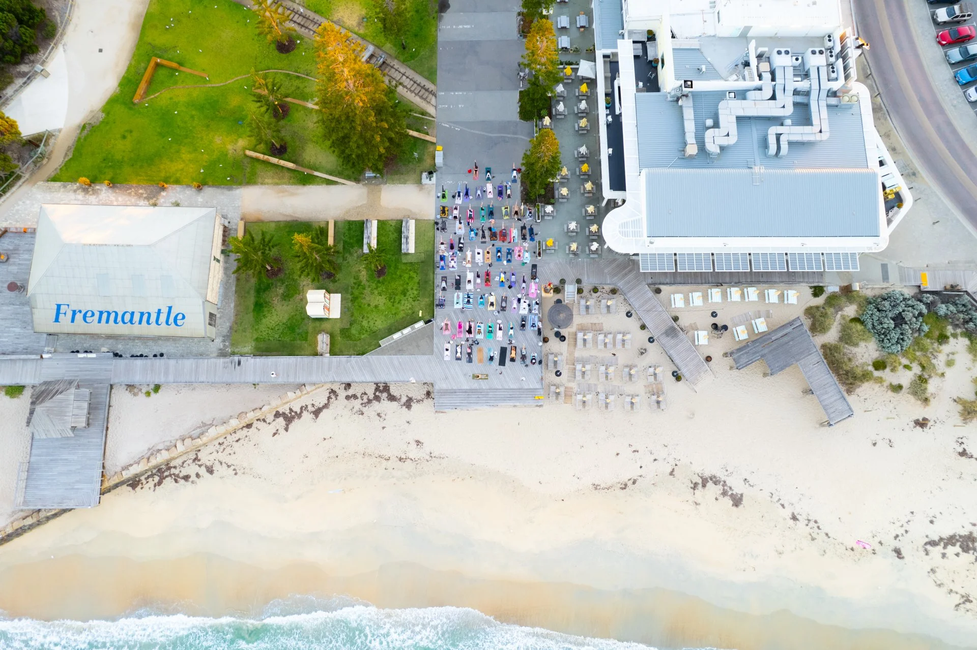 An aerial view of a beach with a boardwalk, grassy park area with trees, a large building with rooftop mechanical units, and a parking lot filled with colorful cars and outdoor seating. The beach has sand and ocean waves.